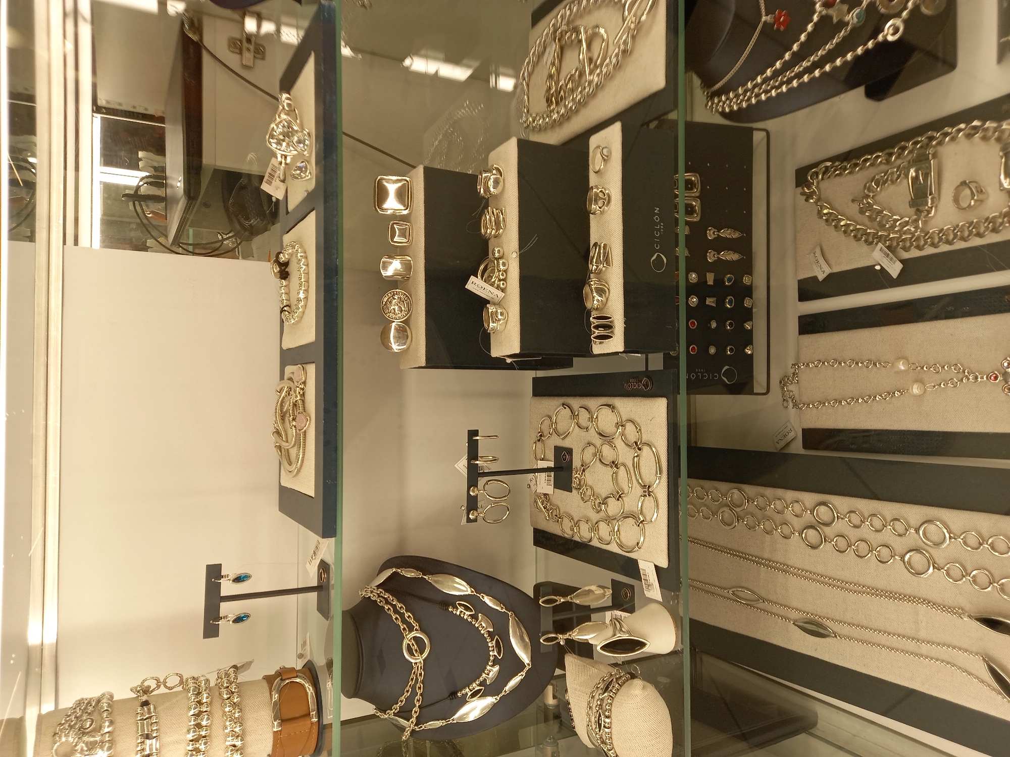 This image shows a jewelry display case in a retail environment. The case contains various pieces of jewelry, including necklaces, bracelets, and earrings, made of gold-toned metal. There are also some silver-toned pieces. The jewelry is arranged on black pedestals and fabric-covered display surfaces. The display case itself is made of glass and is illuminated by overhead lighting. The background appears to be a blank wall with some reflections of lighting fixtures and possibly other store merchandise. There are no people visible in the image. The scene is clean and organized, typical of a jewelry store display. There is no information in the image to suggest a specific location in Yekaterinburg, Russia.