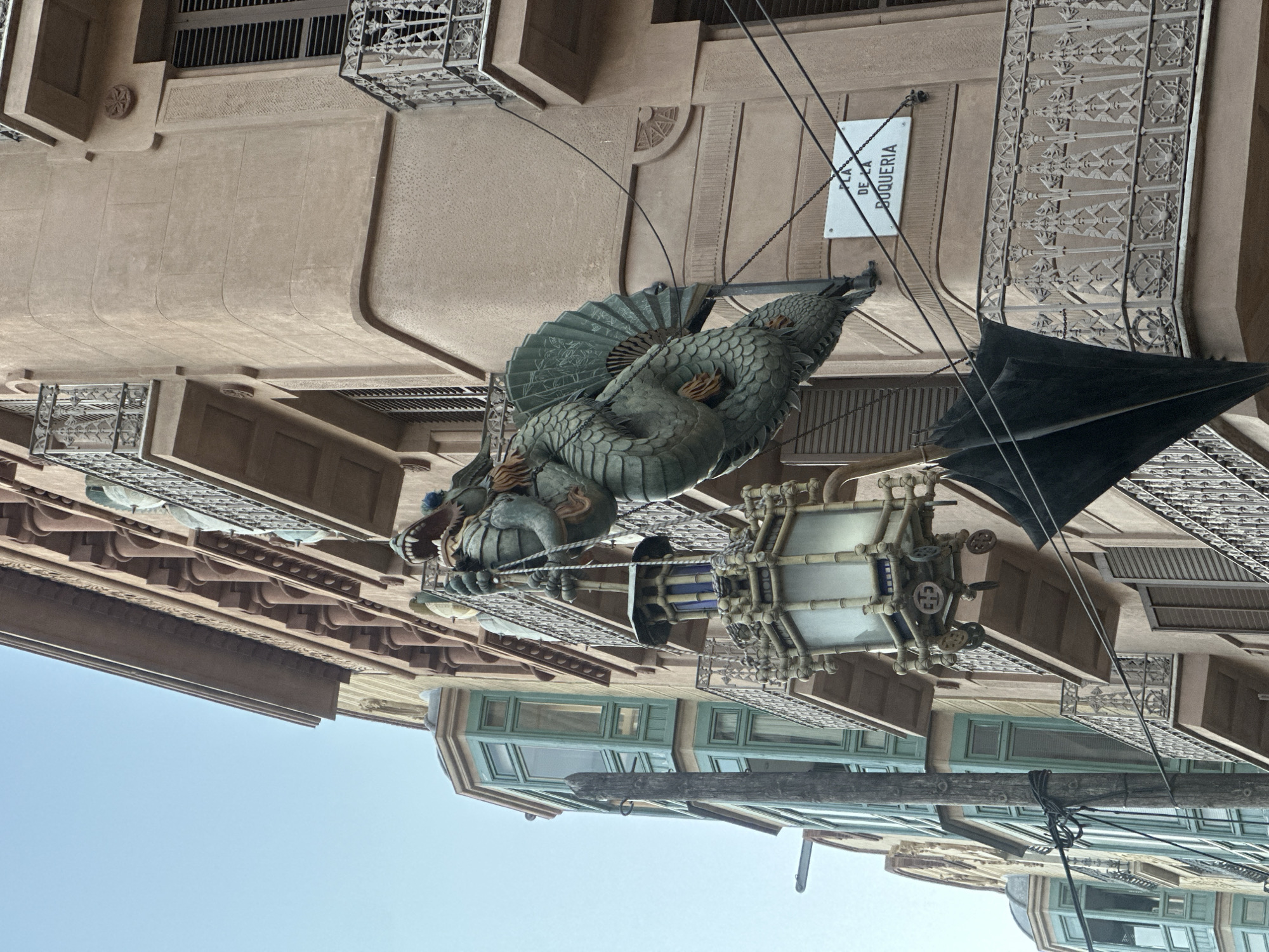 The image captures a street view of a building in Barcelona, Spain, specifically at Plaça de la Boqueria. The building features a distinctive architectural style with balconies adorned with intricate wrought-iron railings. A prominent feature of the scene is a large, green, dragon-like sculpture suspended from the building by chains. The sculpture is positioned above a vintage street lamp, which also appears to be a decorative element. Below the lamp, a dark, umbrella-like object is partially visible. The building's facade is a muted peach color, contrasting with the green of the dragon and the teal window frames of an adjacent building visible on the left. The sky above is clear and pale blue. The image is taken from a low angle, looking upwards at the building and its embellishments.
