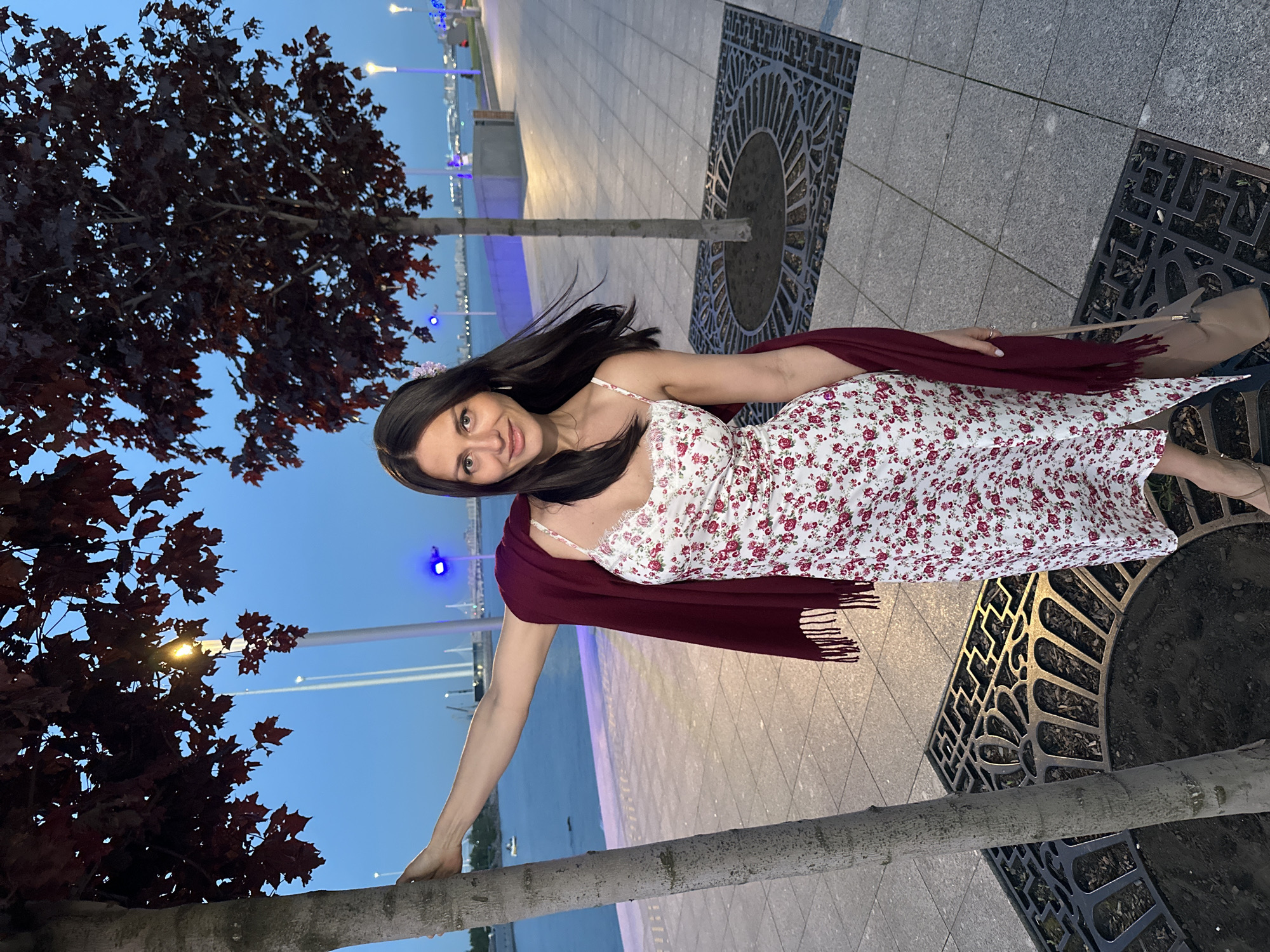 A woman with dark hair stands leaning against a tree in an urban park at dusk. She is wearing a white floral print slip dress with a burgundy shawl draped over her shoulders. The background shows a waterfront promenade with paved walkways, trees, and streetlights. In the distance, a body of water is visible with a city skyline and bridges. The lighting suggests it is early evening, with a clear, blue sky. The scene appears to be a public space in the city of Saint Petersburg, Russia, likely along the Neva River.