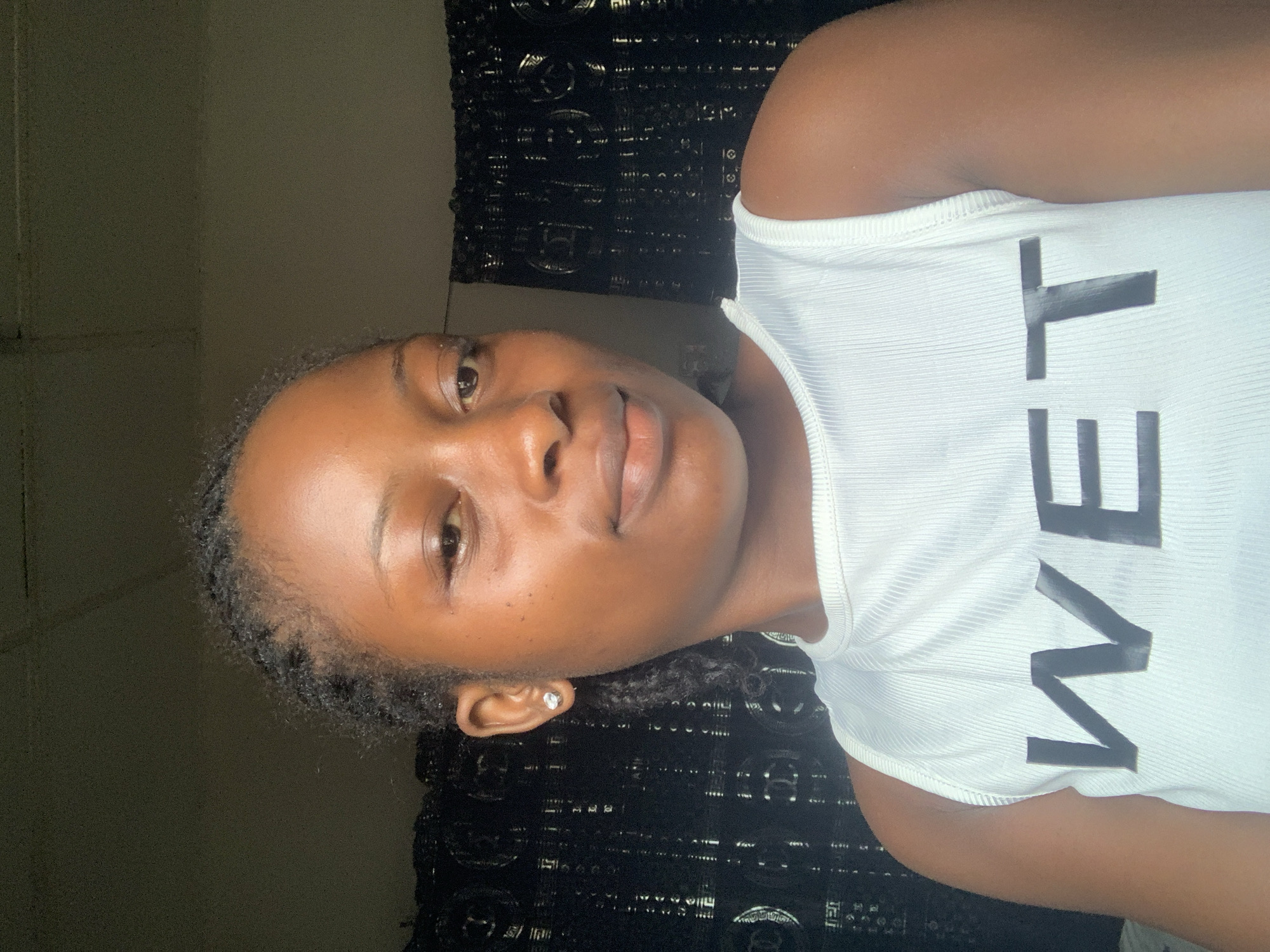 A young, brown-skinned woman is indoors with a black patterned curtain or wall hanging behind her. She is wearing a white tank top with the word "WET" printed in large, black capital letters. She is looking at the camera. Her hair is braided and she is wearing a stud earring in her left ear. The lighting is dim. This photo cannot be used to pinpoint a location in Lokoja.