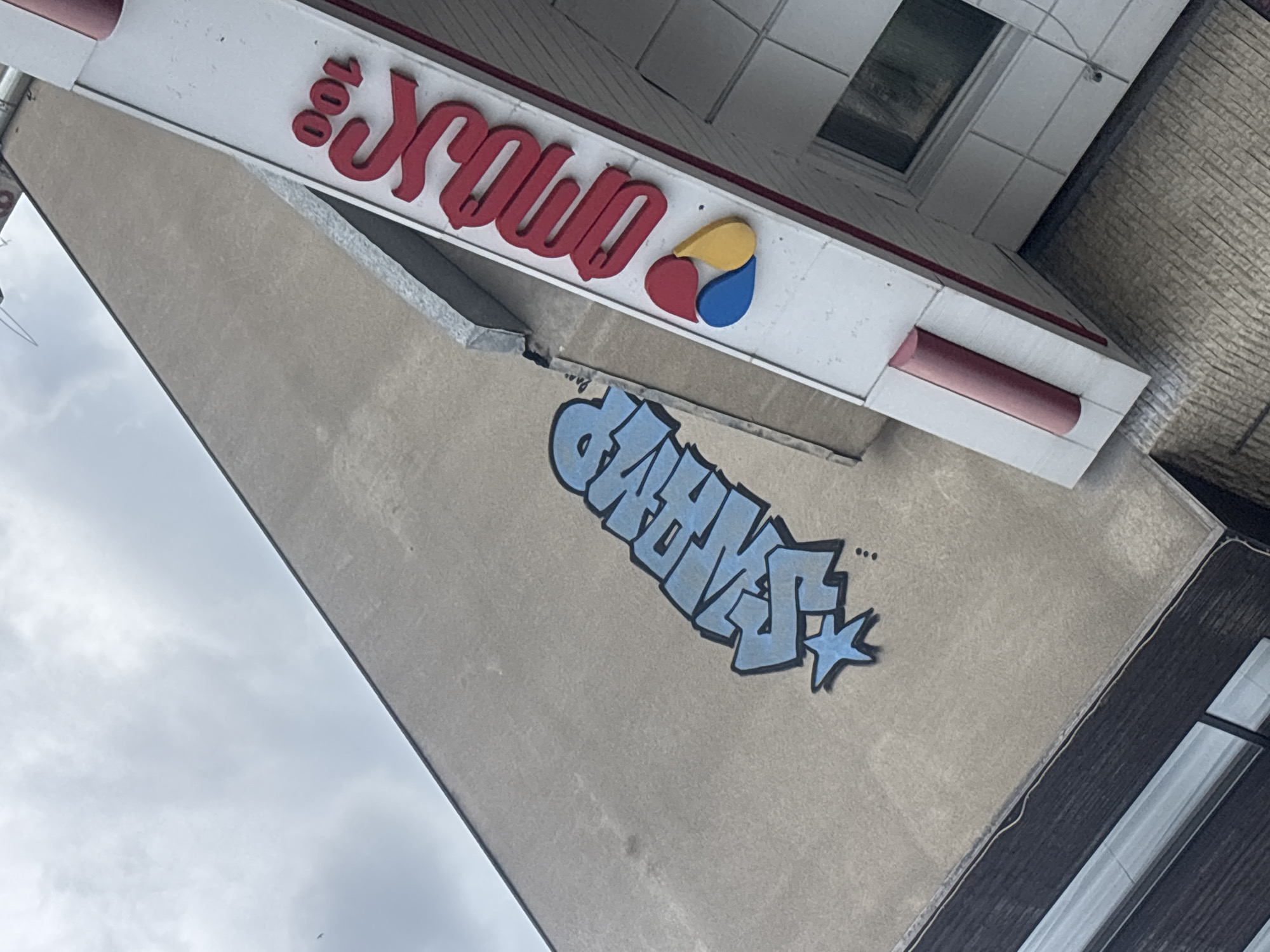 The image depicts the exterior of a building in Tbilisi, Georgia. A sign reading "Krawo 100" in red lettering is prominently displayed. Beneath the sign, graffiti art in shades of blue is visible on the wall. The building's facade is a neutral beige color. The photograph appears to have been taken from a low angle, looking upwards. The sky is cloudy.