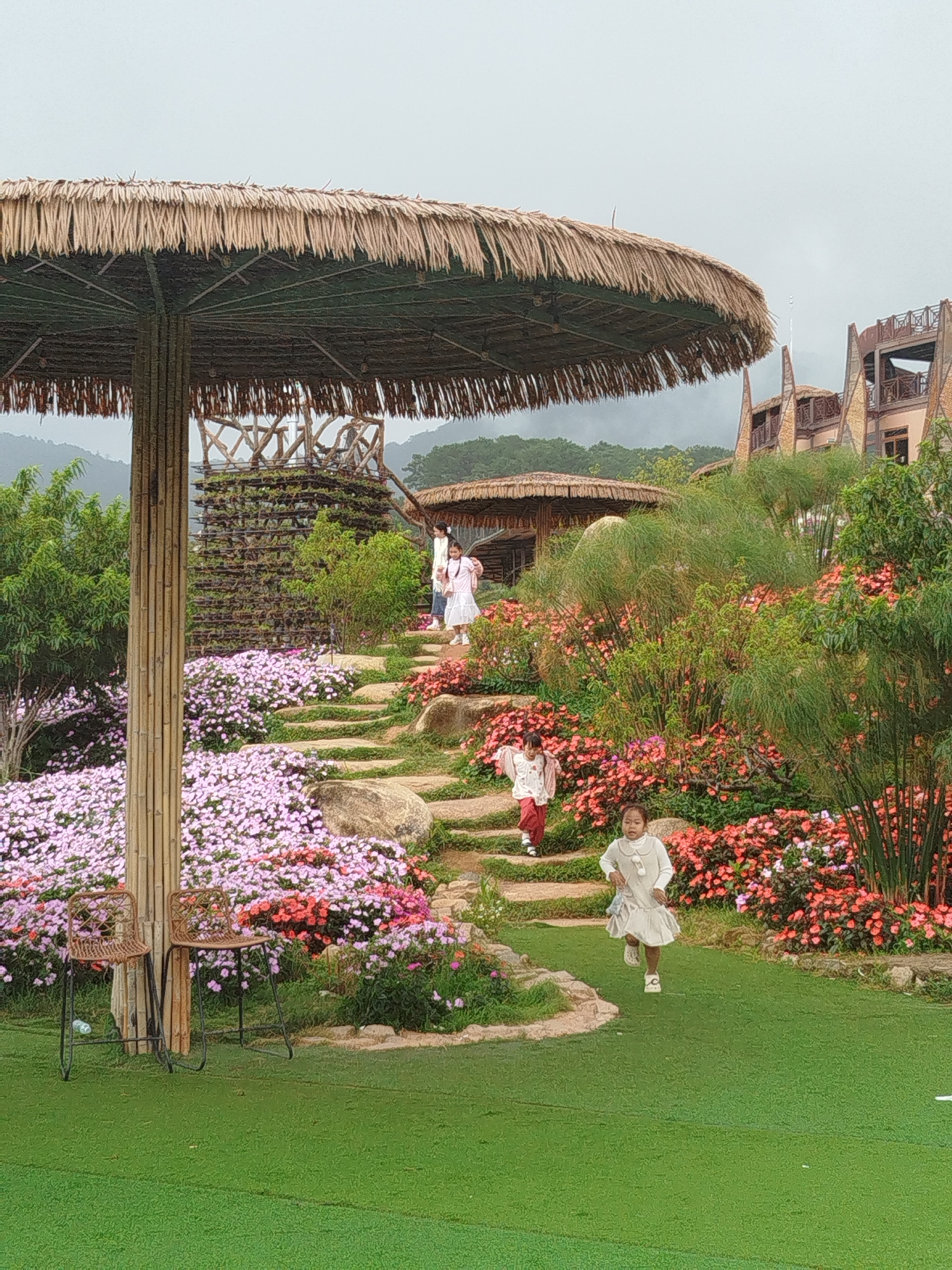 This image captures a scene at a resort or park, likely in Lạc Dương, Vietnam, characterized by lush greenery, vibrant flowers, and an overcast sky. In the foreground, a young girl in a light-colored dress is running across a bright green lawn. Further up a tiered stone pathway, another child in a light-colored outfit is walking. A taller figure, also in a light-colored dress, stands near the top of the stairs. Dominating the left side of the frame is a large, thatched umbrella supported by thick bamboo poles. Beneath the umbrella, two simple brown chairs with metal legs are placed on the lawn. The background features a multi-story building with a distinctive architectural style, partially obscured by trees and vegetation. The landscape is adorned with abundant flowers in shades of pink and coral, arranged along the pathway and bordering the lawn. The overall impression is one of a well-maintained outdoor recreational area, possibly a botanical garden or a tourist attraction.