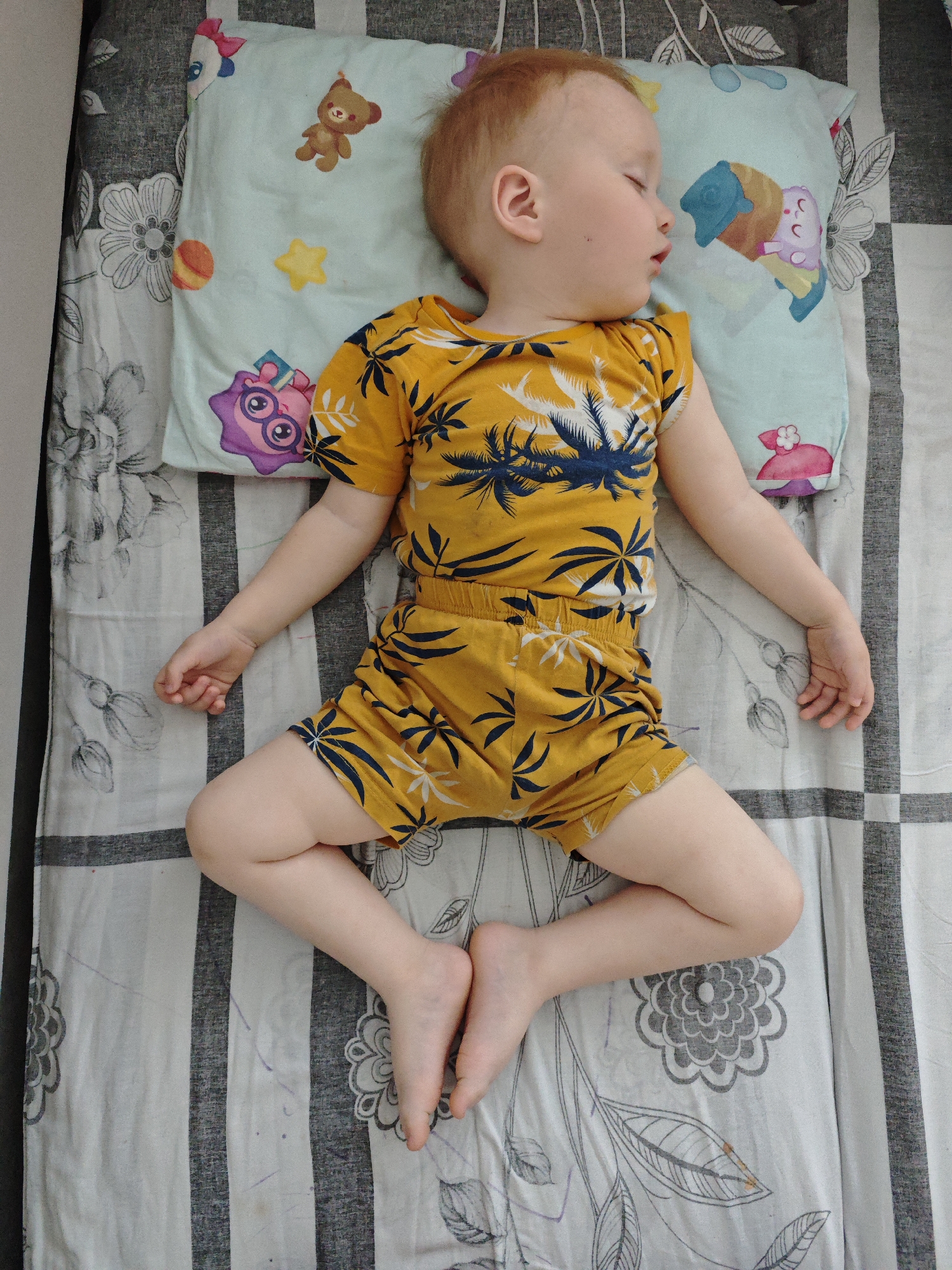 A young child sleeps on a bed in what appears to be an indoor setting. The child is fair-skinned with reddish-blonde hair, wearing a yellow two-piece outfit with a navy-blue palm tree pattern. They are lying on their back with limbs spread out. The child's head rests on a light blue pillow decorated with cartoon-like figures and shapes. The bed covering is patterned with grey and white stripes alternating with floral designs. The image provides no information suggesting that the location is the city of Markovskii, Russia.