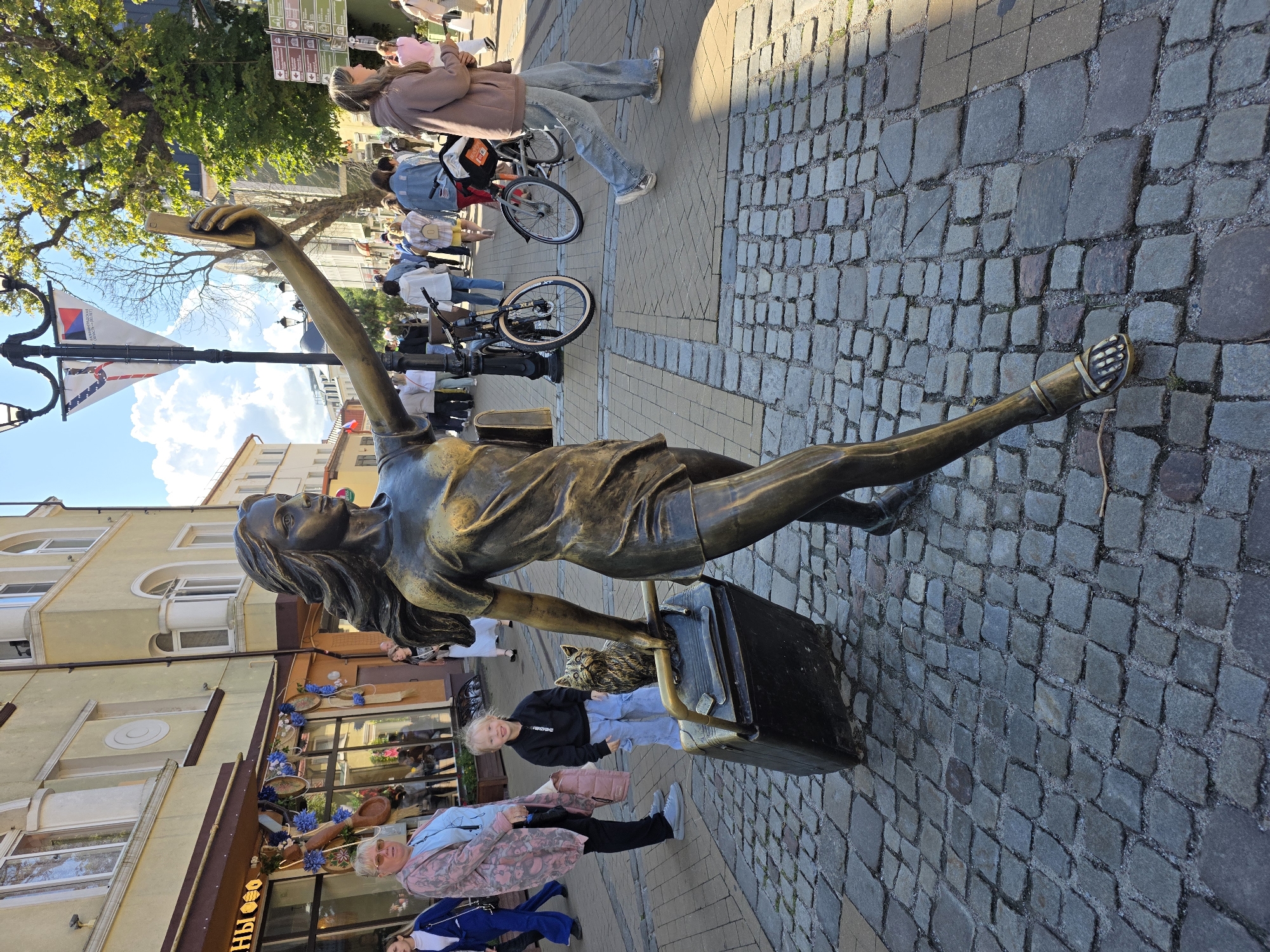 A bronze statue of a young woman is depicted in the foreground, holding a smartphone aloft as if taking a selfie. She is dressed in a short-sleeved, knee-length dress and is pulling a large, rectangular suitcase with her left hand. A small bronze cat sits atop the suitcase. The statue is positioned on a cobblestone street in Zelenogradsk, Russia, a seaside resort town in Kaliningrad Oblast.
To the left of the statue, a woman in a light pink cardigan and sunglasses stands looking towards the camera. Beside her, a young girl with blonde hair smiles at the camera, wearing a black hooded sweatshirt and light blue jeans.
In the background, a bustling street scene unfolds with several people walking and two bicycles parked. Buildings with light-colored facades and arched windows line the street. Trees with green foliage are visible, and a blue sky with white clouds completes the scene. The overall impression is of a lively public space on a sunny day.