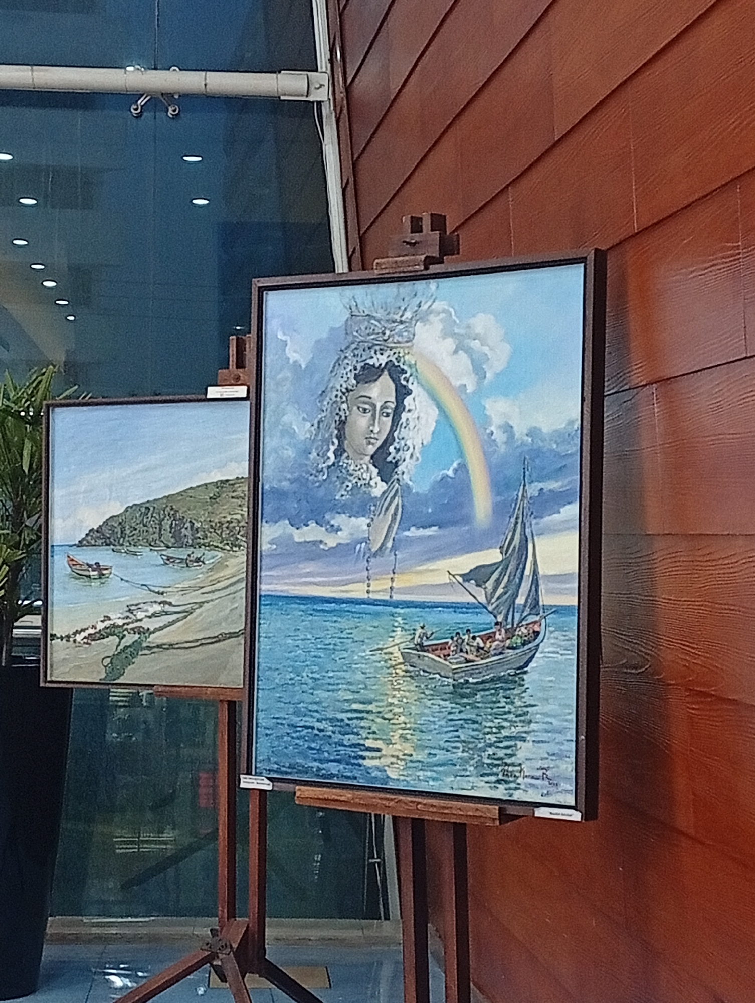 Two paintings are displayed on easels in front of a dark wooden wall. The painting on the left depicts a coastal scene with a rocky hill in the background, several boats in the water near a beach, and clear blue skies. The painting on the right is larger and features a religious theme. It shows a woman with a veil and crown looking downwards, with her hands in prayer. A rainbow arcs across a cloudy sky behind her, and a sailboat with several people on board is visible on the water in the foreground. The environment appears to be indoors, possibly a gallery or exhibition space, with a large glass window or wall behind the paintings. The lighting is soft and diffused.