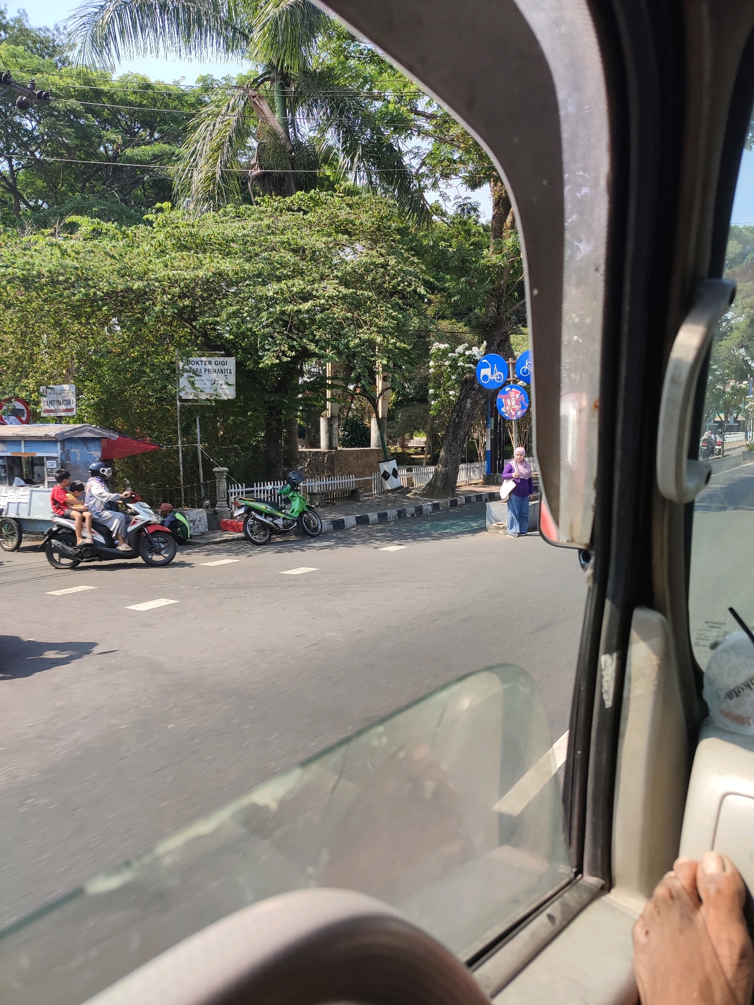 The image, taken from inside a vehicle, shows a street scene in Magelang, Indonesia. In the foreground, a portion of the vehicle's interior, including a steering wheel and a foot, is visible. The view through the window reveals a street with several motorcycles. On the left side of the road, a motorcycle with two young boys is stopped, and a small cart is parked nearby. Further down the road, another green motorcycle is parked.
On the sidewalk, a woman wearing a purple hijab and blue jeans is standing. Behind her, there are trees and signs, one of which appears to be a dental clinic sign. Traffic signs indicating bicycle lanes are also visible. The environment is sunny, suggesting daytime. The road surface is asphalt, and there are pedestrian crossings marked on it. The general impression is of a bustling urban street in Indonesia.