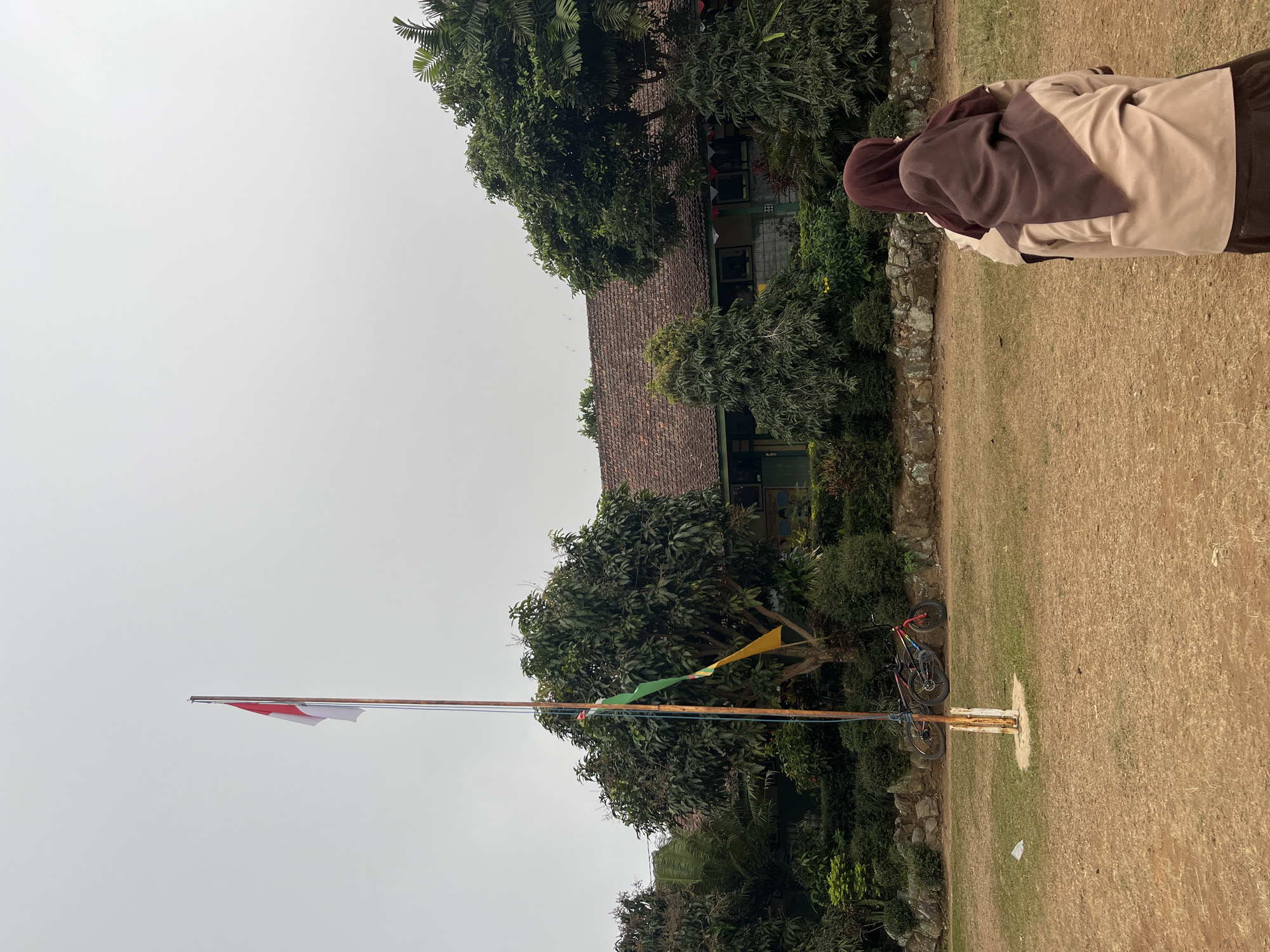 In Gembong, Indonesia, a schoolyard is depicted under a pale, overcast sky. The foreground is a dry, grassy field with patches of dirt. In the mid-ground, a flagpole stands with the Indonesian flag flying at its peak, accompanied by smaller green and yellow flags lower down. Two bicycles are leaning against a low stone wall behind the flagpole. Further back, a single-story building with a tiled roof is visible, partially obscured by lush greenery and trees. Two individuals, seen from behind, are positioned in the right foreground, facing away from the camera and towards the school building. They appear to be students, both wearing light brown uniforms and headscarves, one in a darker brown and the other in a lighter shade. The overall atmosphere is calm and appears to be during daylight hours, possibly before or after a school event.