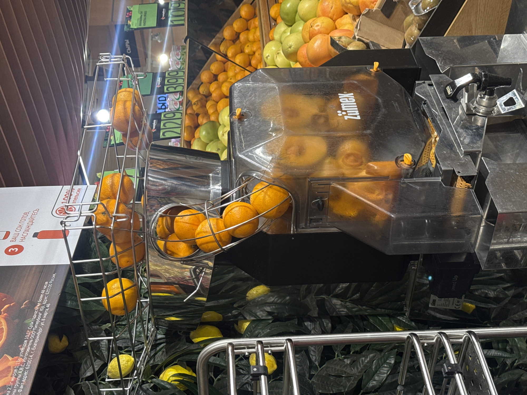 This is a close-up, eye-level, indoor photograph of a commercial citrus juicer in operation. The juicer, identified by the "Zumex" logo, is made of stainless steel and transparent plastic. Several oranges are visible in a metal basket at the top of the machine, and more oranges are being processed or have been juiced, visible within the transparent chamber. A spigot labeled "PRESS" is at the front of the machine, suggesting it's ready to dispense juice. The background shows a display of fresh citrus fruits, including oranges and what appear to be green apples or grapefruits, along with price tags indicating a retail environment. Text on a sign in the upper left corner of the image, partially visible, includes "2,1г жиров", "упаковано 10,1г", and "Екатеринбург, ул. 8 марта, 46". This text identifies the location as Yekaterinburg, Russia, specifically at an address on March 8th Street. The overall scene depicts a food service or grocery store setting focused on fresh juice production.