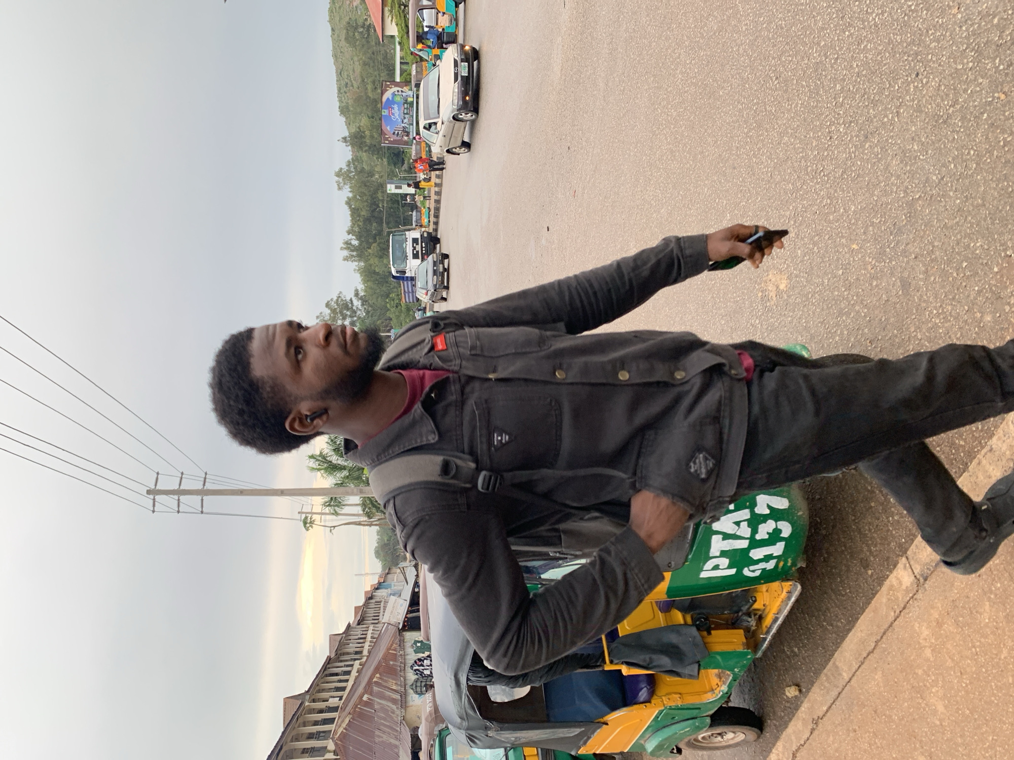 The image depicts a street scene in Gura Top, Nigeria. In the foreground, a young Black man with short, dark, curly hair walks towards the right, his left hand in his pocket and his right hand holding a dark object, possibly a phone. He is wearing a dark denim jacket over a maroon shirt and dark pants, with a dark backpack on his shoulders. To his left, a yellow and green auto-rickshaw, identified by the markings "PTA 1137", is parked on the side of the road. In the background, there are buildings with corrugated metal roofs and balconies, palm trees, utility poles with overhead wires, and other vehicles including cars and a truck. The road surface appears wet, suggesting recent rain. The sky is overcast.