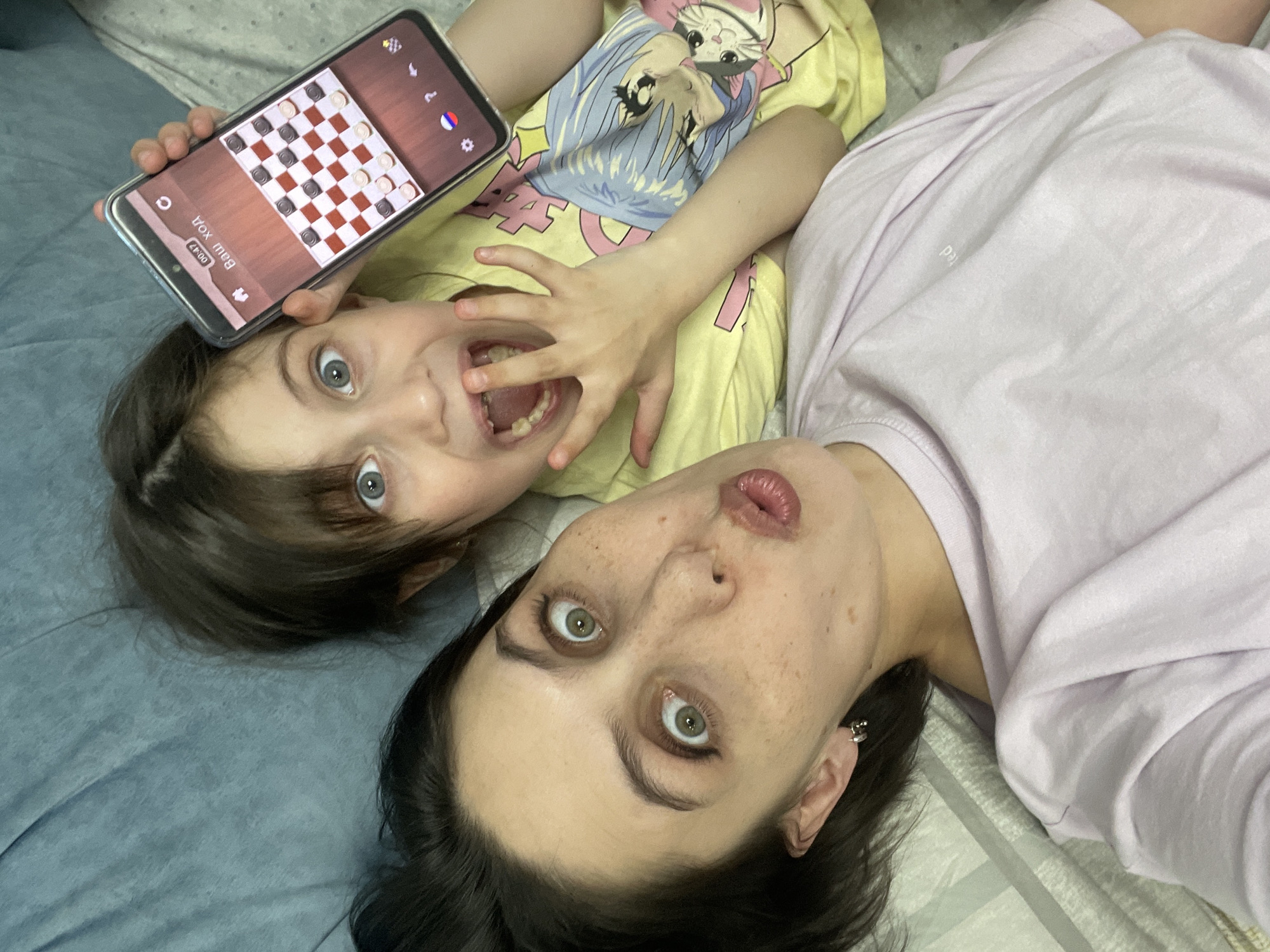 A selfie-style shot depicts a child and a woman lying on a blue and white patterned surface, potentially a bed or bedding. The child, wearing a yellow t-shirt with a cartoon print, is positioned with a mobile phone resting above their head. The phone screen displays a checkers game. The child's mouth is open, and their hand is near their face. The woman, in a lavender shirt, lies next to the child. Her expression includes puckered lips. Her dark hair contrasts with her pale skin. There is no evidence to confirm that this photograph was taken in Izhevsk, Russia.