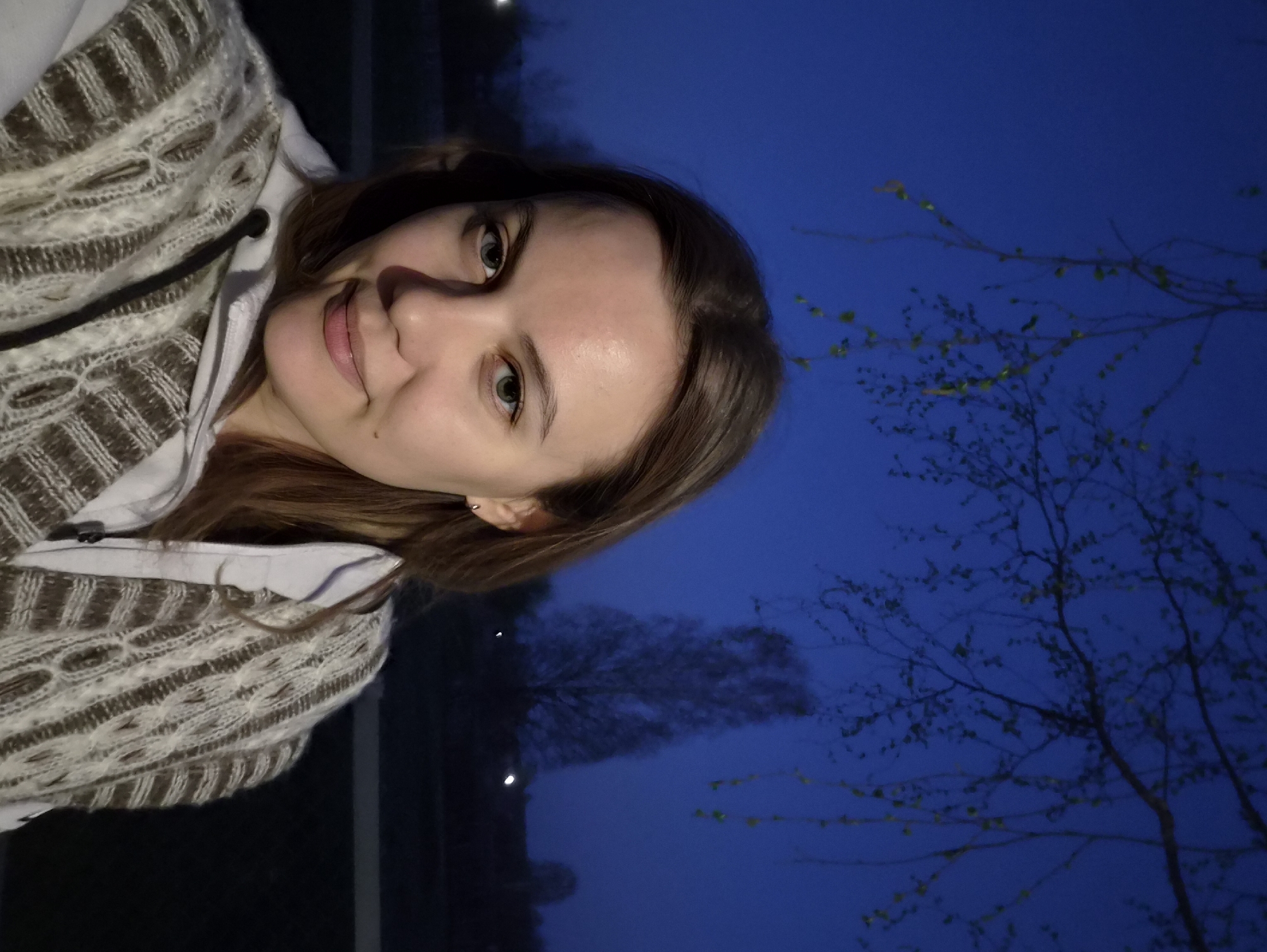 A selfie shows a woman with light skin, brown hair, and green eyes, wearing a brown and white sweater over a white shirt. She is smiling slightly. Behind her is a dark blue sky with trees silhouetted against it, with branches displaying new spring leaves. Two small lights are visible in the distance. It is not possible to determine the exact location of the photo, even given the city is Узяр, Russia.
