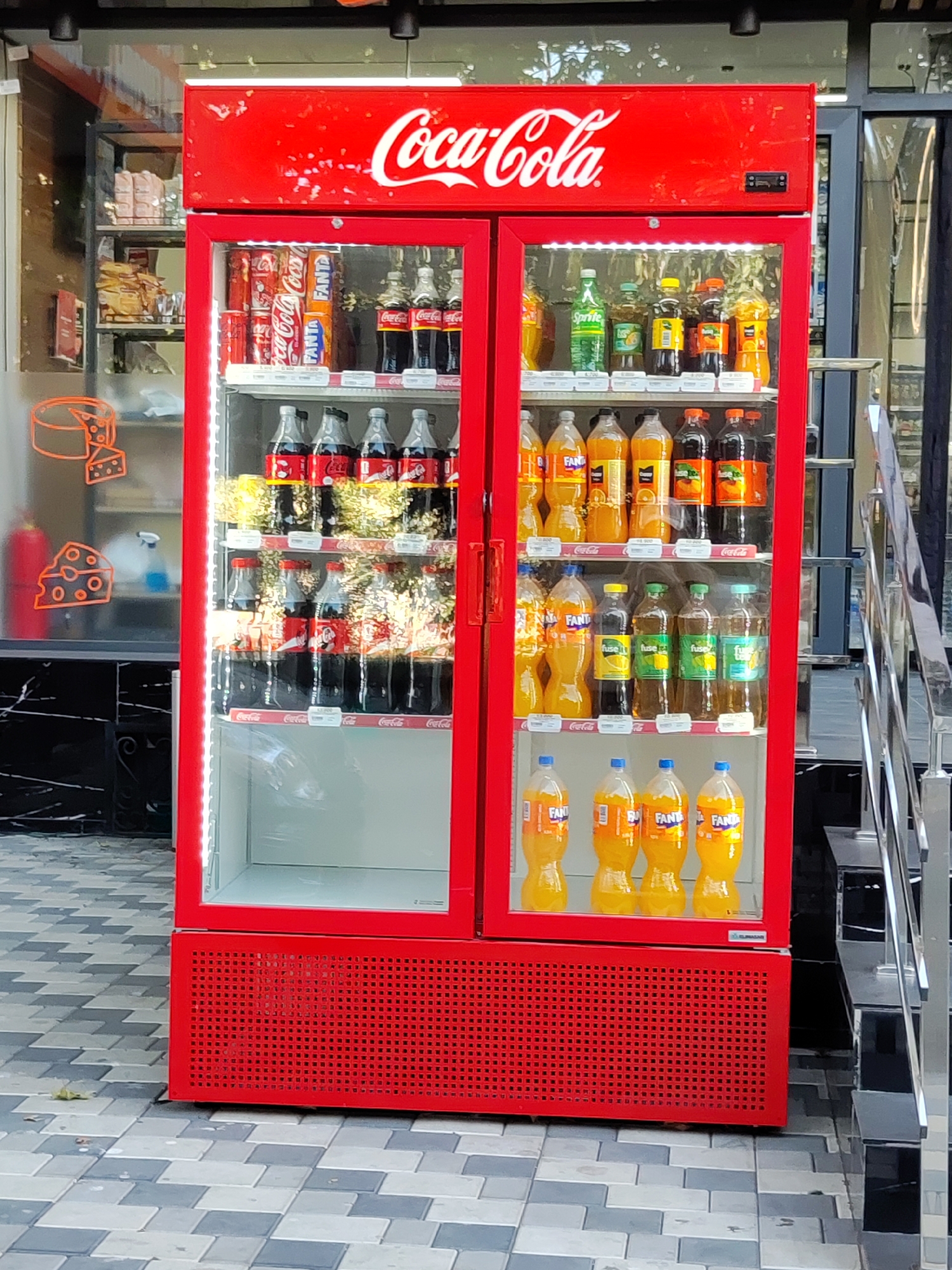 An outdoor Coca-Cola branded refrigerator is filled with various bottled beverages and cans. The refrigerator is bright red with a transparent front, allowing clear visibility of the products inside. The top of the refrigerator displays the iconic "Coca-Cola" logo in white lettering.
The refrigerator is divided into two sections, each with multiple shelves. The left section contains cans of Coca-Cola and Fanta, along with large bottles of Coca-Cola. The right section is stocked with a variety of bottled drinks, including Fanta, Sprite, and other assorted beverages in different colored bottles. The bottles are arranged in neat rows on each shelf. Some shelves have price tags visible.
The refrigerator is situated on a paved walkway made of grey and white square tiles, laid in a patterned arrangement. To the right of the refrigerator, a metal staircase with a silver railing is visible. In the background, beyond the refrigerator, there is a store entrance with glass doors and windows, and some products are visible on shelves inside the store, including what appears to be cheese illustrations on the glass. The scene appears to be an exterior of a shop or convenience store in the city of Tashkent, Uzbekistan. There are no people visible in the image.
