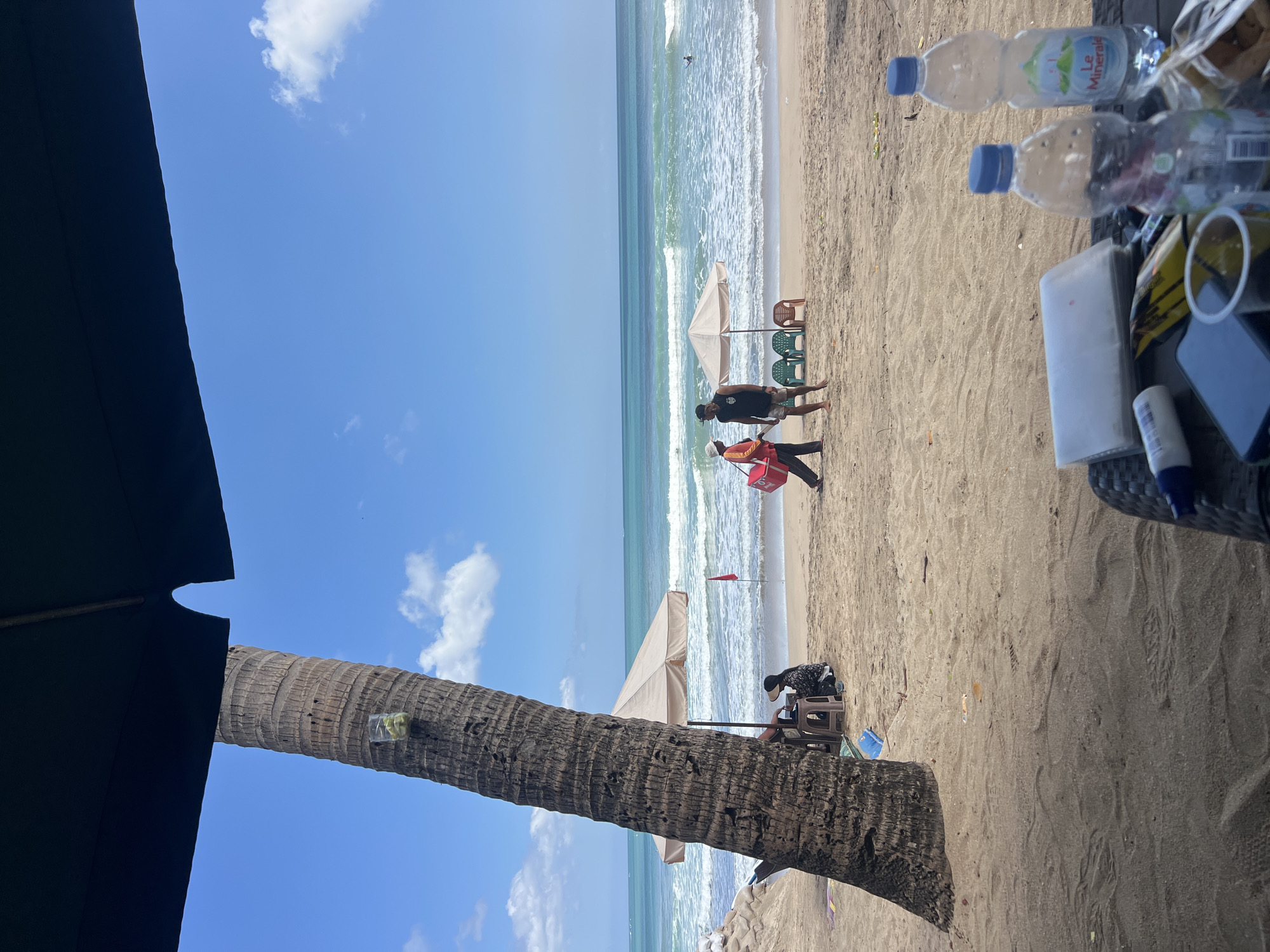 The image shows a beach scene in Kuta, Indonesia. A large palm tree dominates the left side of the frame, with a dark umbrella extending over the top. In the background, the turquoise ocean meets a sandy beach where several people are present. Two individuals are walking near a beach umbrella and chairs. Another person is sitting under an umbrella with a red flag planted in the sand nearby. In the foreground, a table holds two plastic water bottles, a phone, a plastic cup, and some other items. The sky is blue with scattered white clouds.