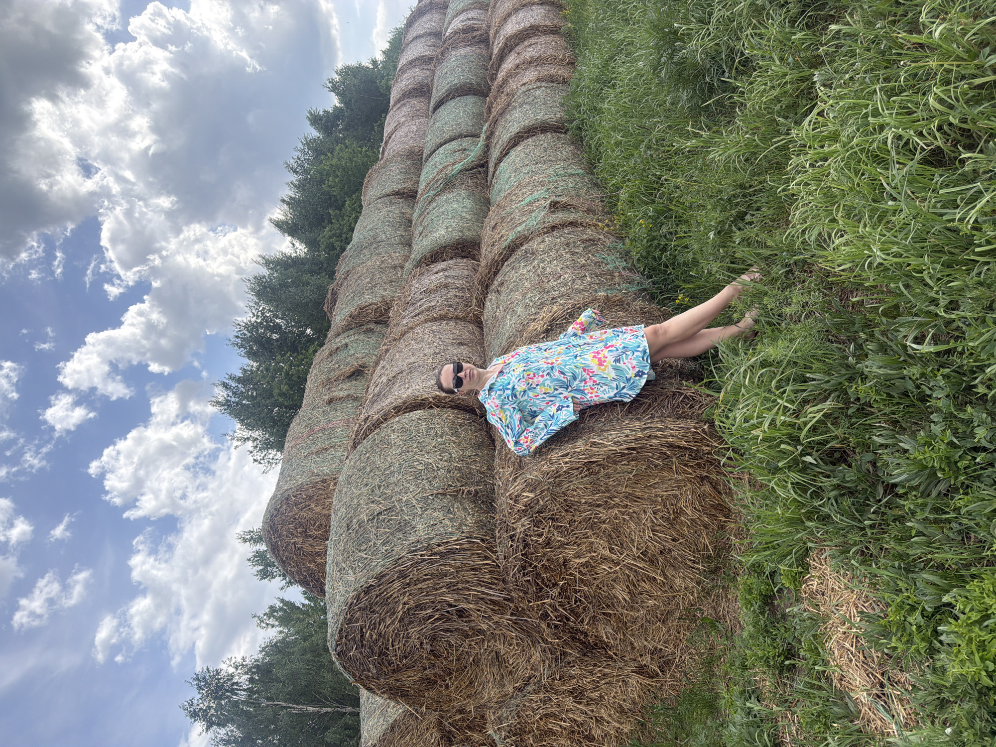 A woman is lying on a stack of hay bales in a grassy field. She is wearing a floral dress and sunglasses. The hay bales are stacked high, reaching towards the sky. The sky is partially cloudy. The location is Kirillovka, Russia.