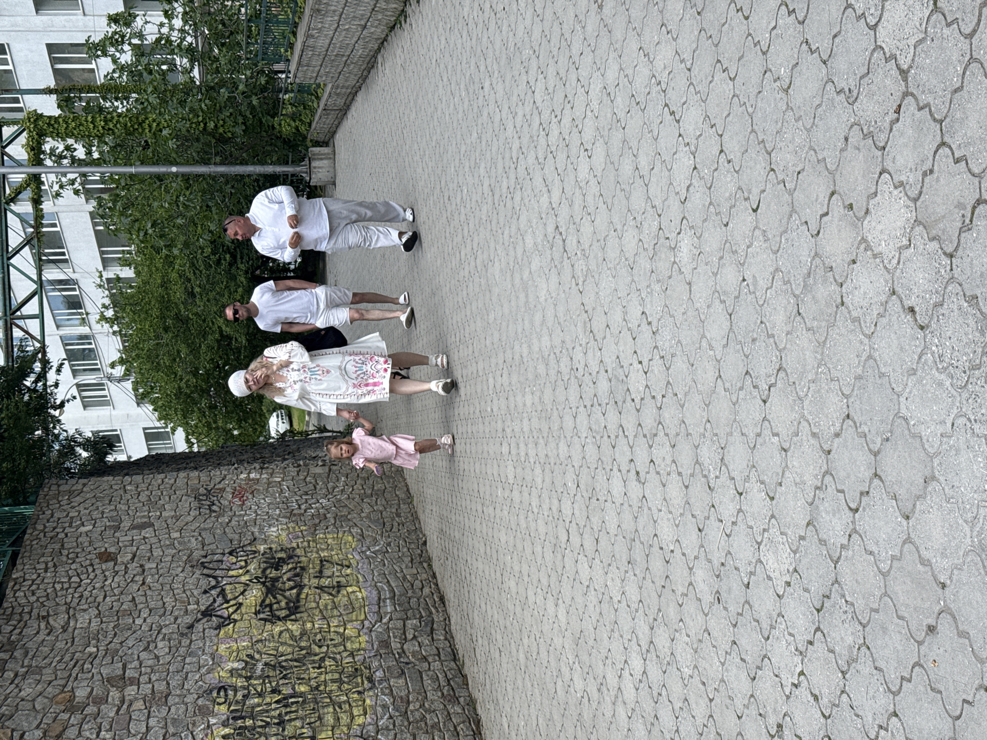 The image depicts four individuals walking on a patterned stone walkway next to a stone wall covered in graffiti. The person closest to the camera is a young girl in a pink dress and light-colored shoes. Behind her is a woman wearing a white dress with embroidery, a white cap, and white shoes. Further along are two men, both wearing white shirts and light-colored shorts or pants. One is wearing sunglasses and white shoes, while the other is wearing darker shoes. The background includes greenery, a building with windows, and a metal structure. The location appears to be an urban environment, potentially a park or pedestrian area.
