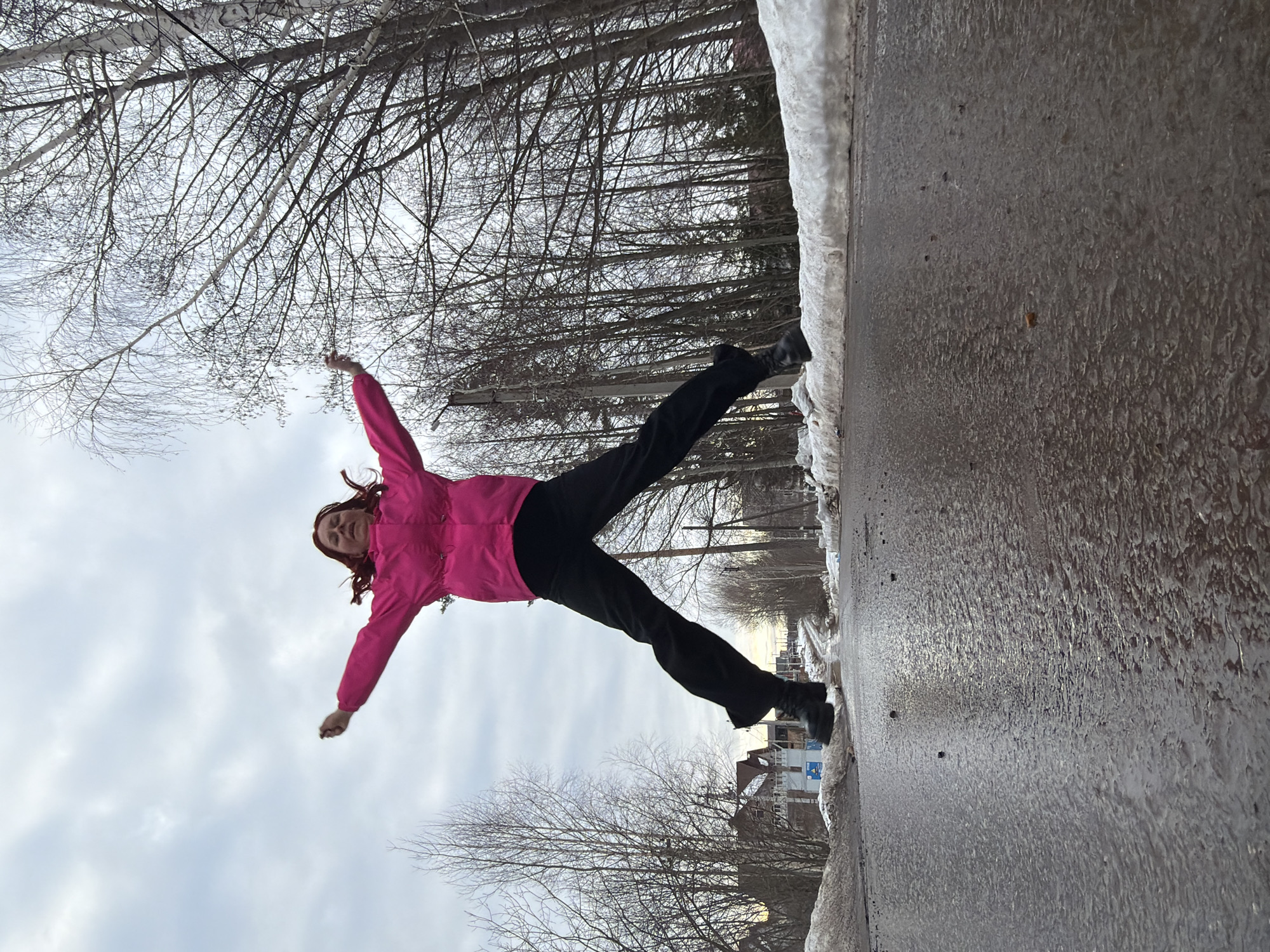 A single individual, a person with reddish-brown hair, is captured mid-air, jumping with arms outstretched and legs splayed wide, wearing a bright pink hooded jacket, black pants, and black boots. The scene is an outdoor setting on a wet, dark asphalt road, indicative of melting snow or recent precipitation. Banks of melting snow line both sides of the road. Bare deciduous trees, including birch, frame the road on both the left and right, with visible utility poles on the right. In the distant background on the left, residential buildings are visible, including one with a reddish-brown roof and another under construction with blue coverings. The sky is overcast with scattered clouds. The image is taken from a low-angle perspective, looking up towards the subject and the sky. The location is in the city of Chaikovsky, Russia.