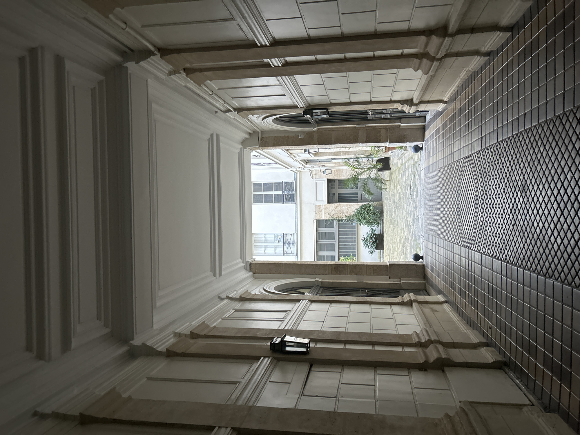 The image depicts a courtyard viewed from a low angle looking upwards. The courtyard is enclosed by buildings with white facades and windows, and is accessible through a covered walkway. The walkway has a tiled floor on the right side and white paneling on the left. The ceiling of the walkway is divided into rectangular panels. At the end of the walkway, greenery and a glimpse of the sky are visible. A light fixture is mounted on one of the panels on the walkway ceiling. The location is in a city, but the specific city and country are unknown.