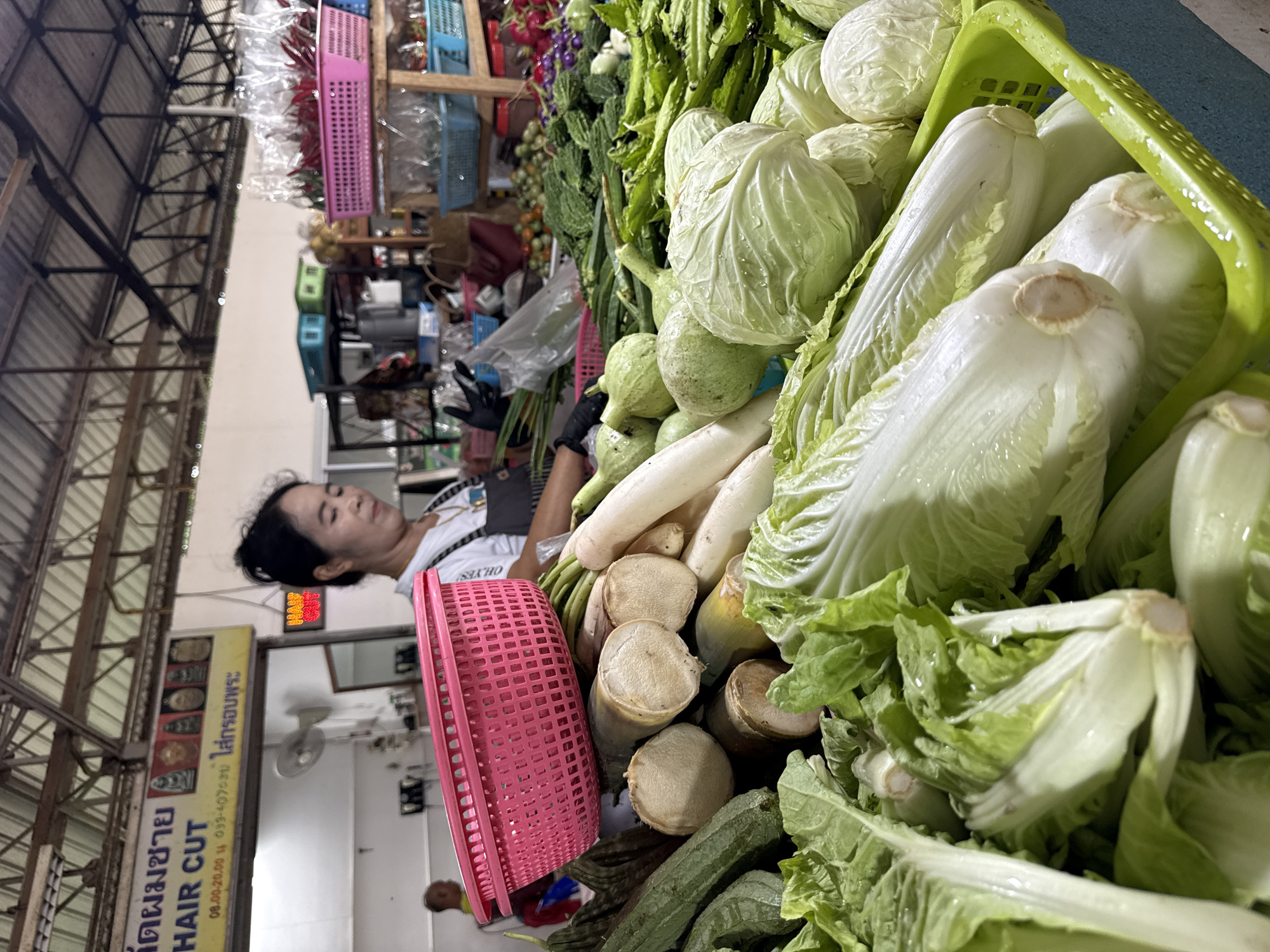 This media file captures a woman working at a market stall filled with fresh vegetables in Baan Bo Phut, Thailand. The scene is oriented vertically. The woman, wearing a white t-shirt and black gloves, is leaning over, appearing tired or focused on her task. In the foreground, a large assortment of vegetables is displayed, including cabbage, daikon radishes, gourds, and leafy greens, some of which are wet, suggesting they have recently been washed or are dewy. A pink basket is placed among the vegetables. Behind the woman, a sign with Thai text and the English words "HAIR CUT" is visible, indicating a nearby business. Above the sign, a digital display shows numbers. The background reveals market structures, including metal beams and roofing, as well as shelves stocked with items in colored baskets and plastic packaging. The overall impression is a busy marketplace scene, likely during daylight hours, given the lighting.