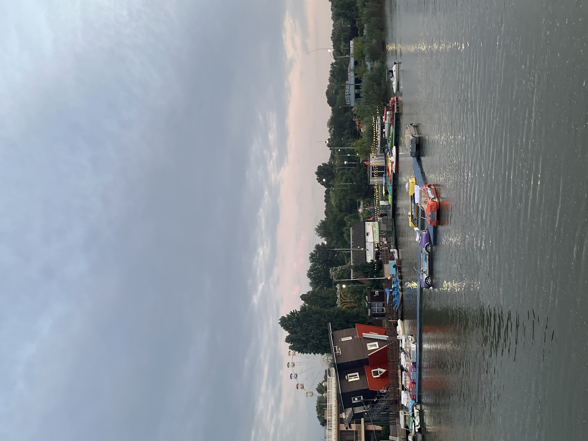 The image depicts a waterfront scene in Krasnodar, Russia, likely at an amusement park or recreational area. In the foreground, a calm body of water reflects the overcast sky and surrounding lights. Several small, colorful pedal boats, designed to resemble cars, are docked or cruising on the water. One red pedal boat with "ELECTRO" written on its side is prominently featured. In the middle ground, a long blue platform hosts a variety of other pedal boats and small craft. Adjacent to this, a peculiar attraction stands out: a building, seemingly a house, is constructed upside down, with its foundation pointing towards the sky. Behind the upside-down house, a Ferris wheel with colorful cabins is visible, along with trees and other structures. The overall atmosphere suggests a leisurely and perhaps slightly whimsical environment. The sky is a mix of grey and soft pink hues, indicating it may be dusk or dawn.