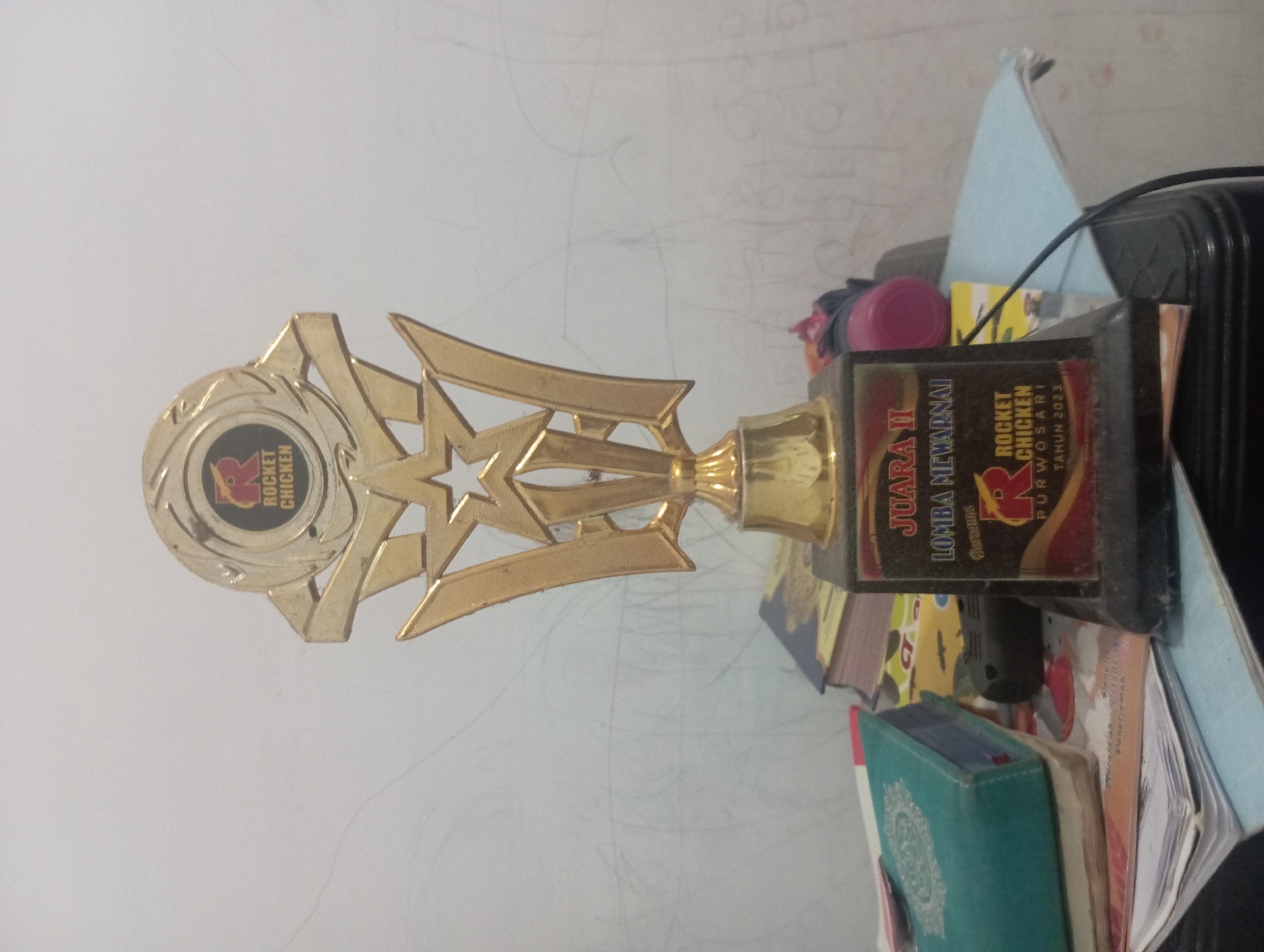 The image shows a trophy placed on a table. The trophy is gold and black and has text that reads "JUARA II LOMBA MEWARNAI ROCKET CHICKEN PURWOSARI TAHUN 2023". There are several books and papers on the table, and a wall with faint drawings in the background. The trophy appears to be a prize for a children's coloring competition organized by Rocket Chicken in Purwosari, Indonesia, in 2023. The overall scene suggests a casual indoor setting, possibly a home or office. There are no people or animals visible in the image.