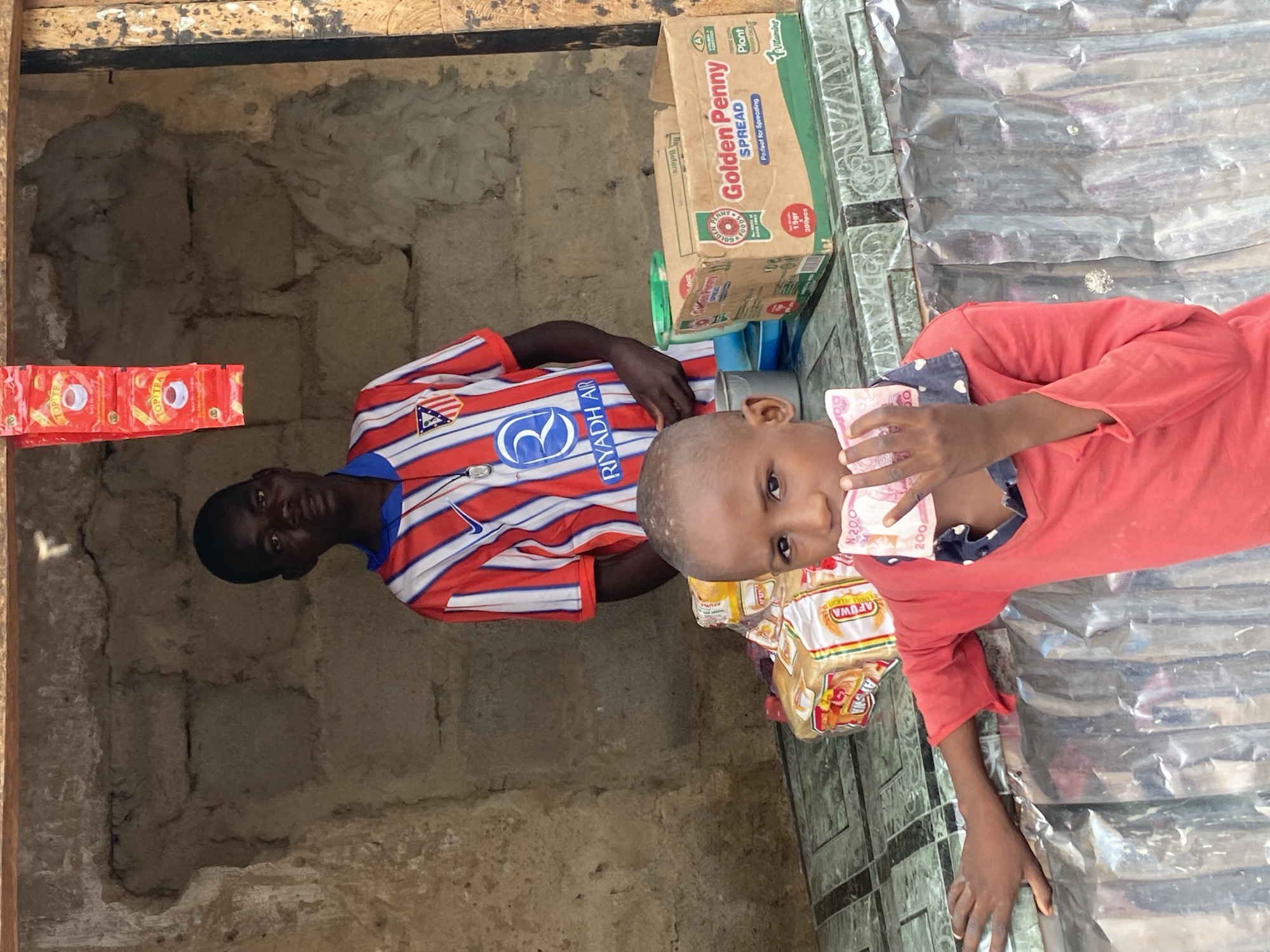 A medium shot shows two young boys in front of a market stall in Rigachikun, Nigeria. The boy in the foreground, wearing a red shirt with a dark blue collar, holds a wad of Nigerian Naira bills to his mouth. He has short, dark hair and is looking directly at the camera. Behind him, a slightly older boy, wearing a red, white, and blue striped athletic shirt with "RIYADH AIR" printed on it, stands in the stall. The stall is constructed of rough, light-colored bricks and has a wooden frame at the top. Red packets of tea are hanging from the top of the stall. Several loaves of bread wrapped in plastic are visible on the counter, along with a box labeled "Golden Penny SPREAD." The counter surface appears to be tiled in a green and grey pattern. The background is a rough, unpainted brick wall, and the foreground features a corrugated metal surface.