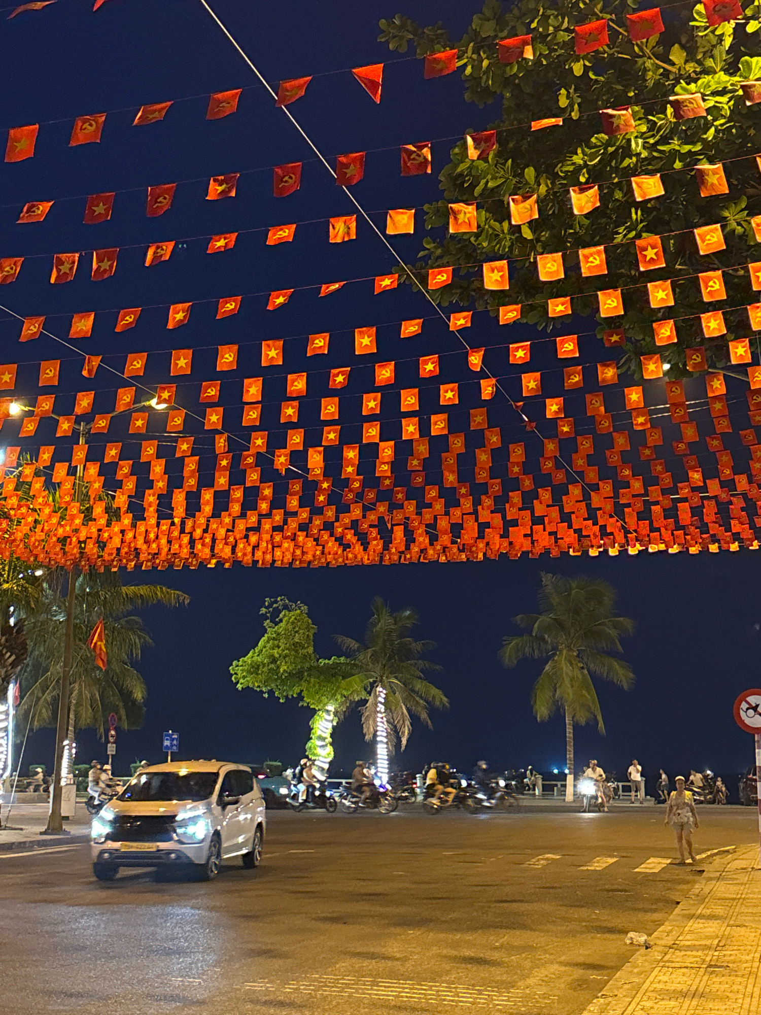 The image shows a nighttime street scene in Nha Trang, Vietnam, likely along Tran Phu Street. A white SUV is driving on the road, its headlights on, facing the camera. The street is lined with palm trees, and overhead, numerous red and yellow triangular flags, some featuring communist symbols like the hammer and sickle and stars, are strung across the street, likely for a celebration or holiday. In the background, there are other vehicles, including motorbikes, and people walking along a waterfront promenade. The sky is dark, indicating it is nighttime.