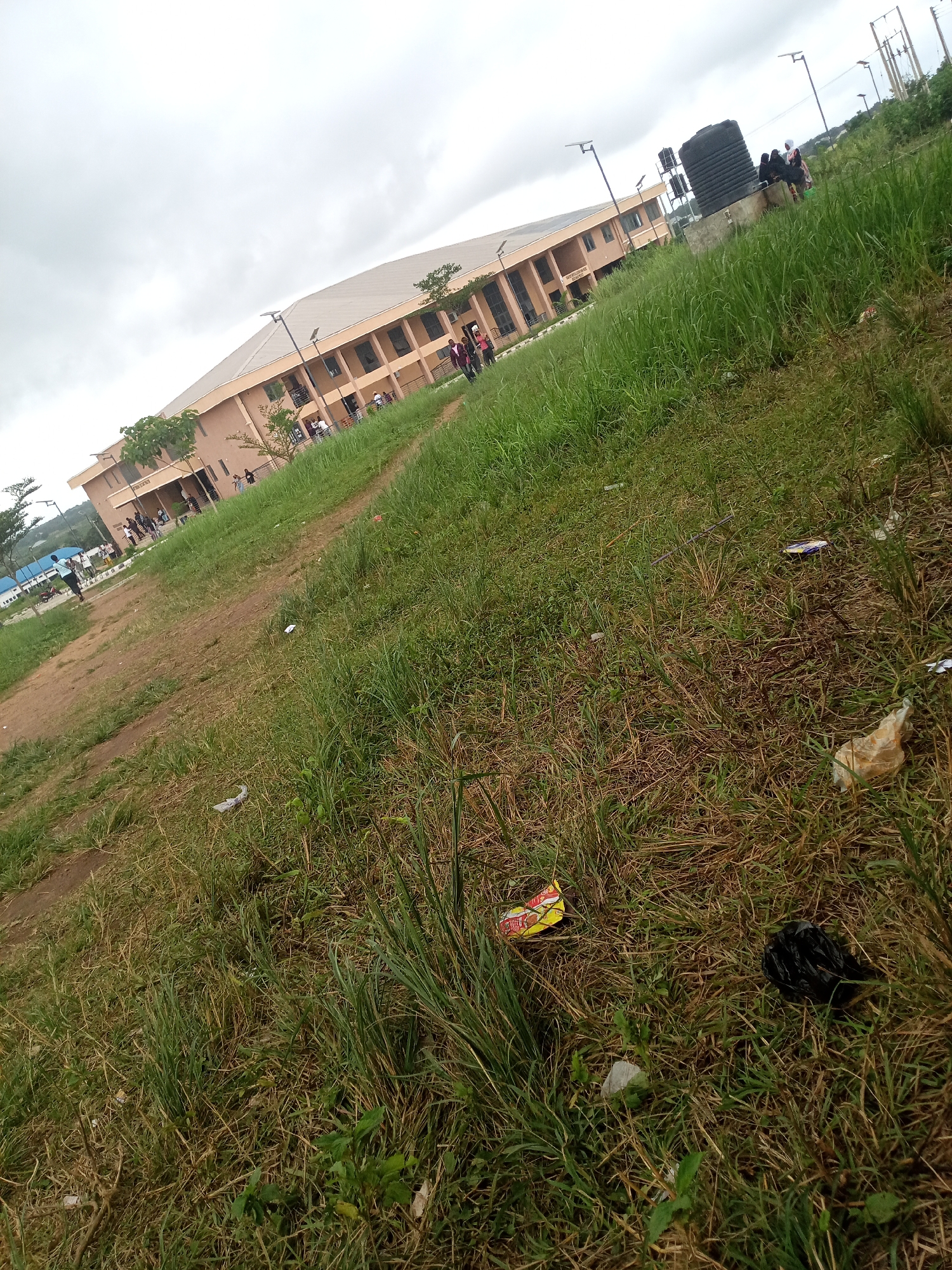 The image depicts an outdoor scene on an overcast day, likely at a university or institutional campus in Iseyin, Nigeria. In the background, a tan, multi-story building with a sloped roof and visible windows is prominent. To the left of the building, a pathway or dirt road is visible, with a few individuals walking on it. In the middle ground, tall green grass dominates the foreground, with a large black water tank situated amidst the vegetation. Several streetlights are also visible throughout the scene. The ground in the foreground is littered with various pieces of trash, including plastic bags, wrappers, and other debris, suggesting a lack of cleanliness in the immediate area.