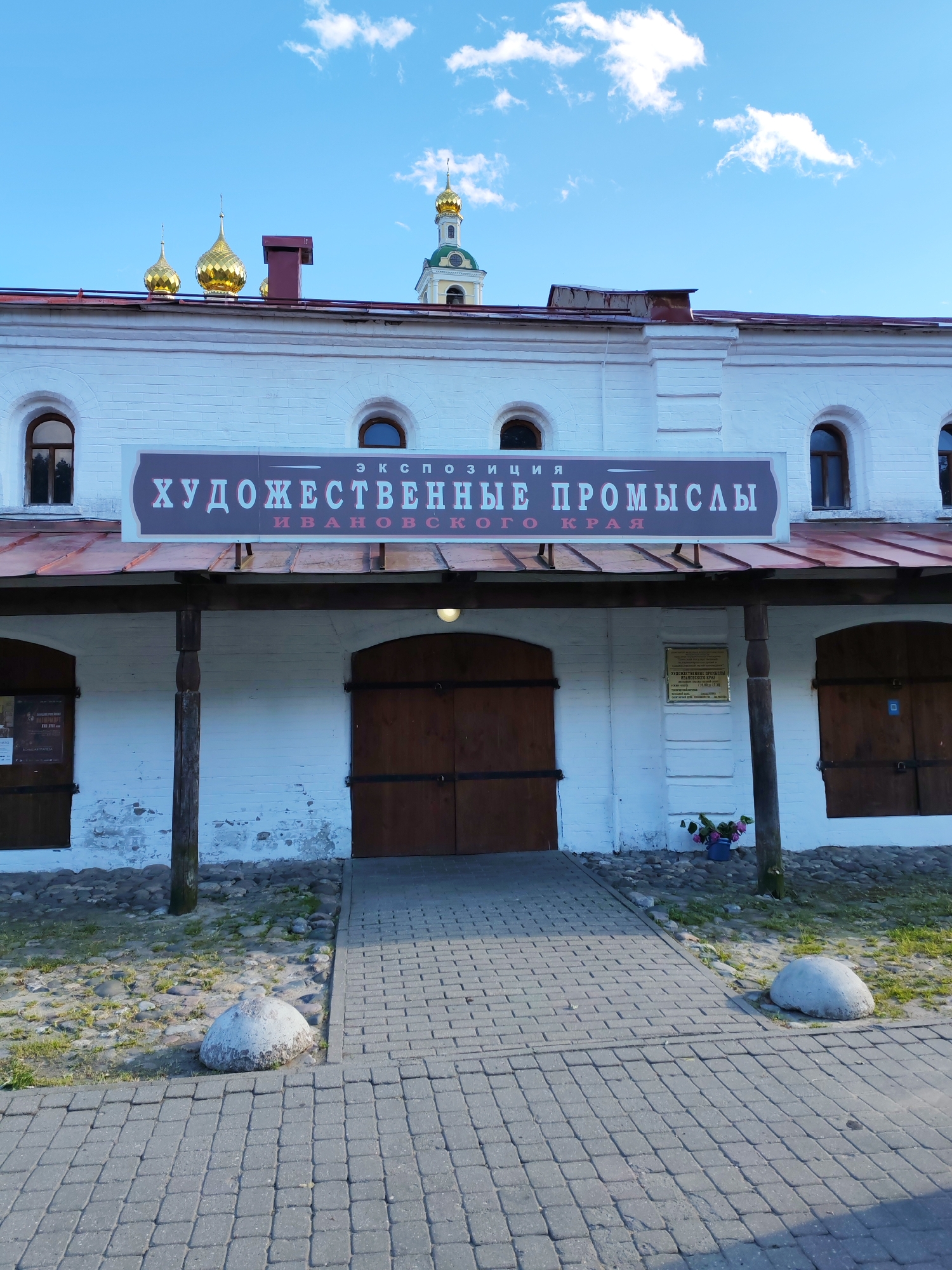 The image shows the exterior of a building with a sign that reads "ЭКСПОЗИЦИЯ ХУДОЖЕСТВЕННЫЕ ПРОМЫСЛЫ ИВАНОВСКОГО КРАЯ" (EXHIBITION ARTISTIC CRAFTS OF IVANOVO REGION). The building is white with arched windows and large wooden doors. A covered walkway with wooden posts runs along the front of the building. Above the building, golden domes of a church are visible against a bright blue sky with white clouds, suggesting a sunny day. The foreground shows a paved walkway leading to the building, with cobblestones and grassy areas on either side. Large, rounded stones are placed along the edge of the walkway. A sign is mounted on the wall to the right of the entrance. The overall scene appears to be an outdoor view of a historical or cultural institution, possibly a museum or exhibition hall, in Plyos, Russia. The time of day is likely daytime, given the bright lighting.