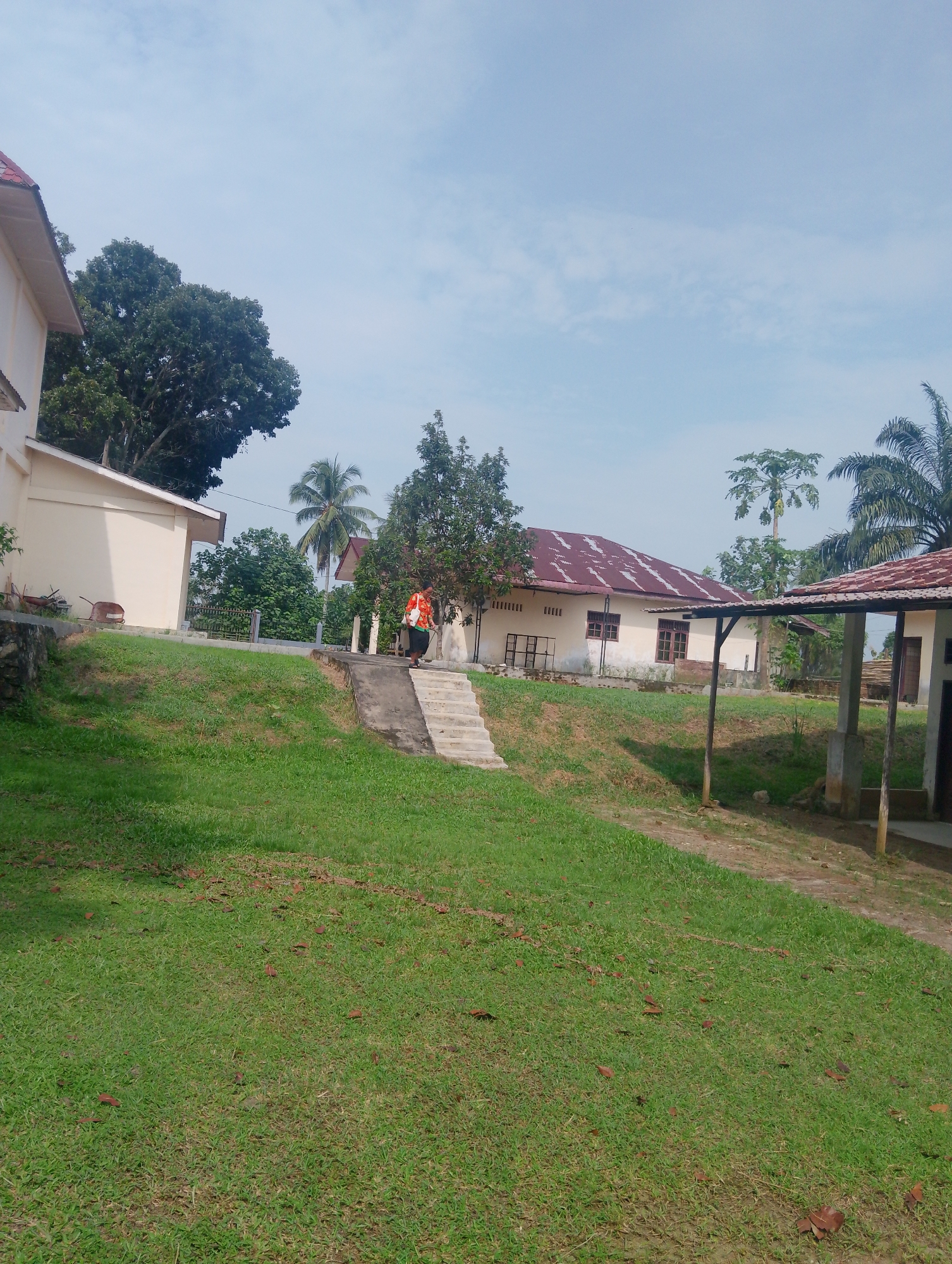 A full outdoor shot shows a rural scene in Serbelawan, Indonesia. The sky is a pale blue with scattered clouds. In the foreground, a lush green lawn slopes upward. A concrete path with stairs runs up the side of the lawn, leading to a building with a dark tiled roof and cream-colored walls. Two individuals, possibly school children based on their small size and bright orange vests, are seen climbing the stairs. Behind them, another cream-colored building with a similar roof sits on the left side of the frame. A tall palm tree is visible in the background, along with dense green foliage and more buildings, suggesting a small community or school campus. The overall impression is one of a sunny day in a tropical, rural environment.