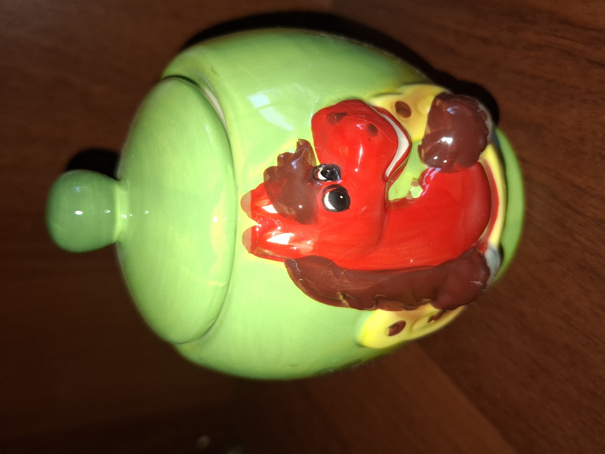 A bright green ceramic jar with a lid sits on a brown wooden surface. The front of the jar features a raised, red, cartoonish horse head with dark brown mane and ears. The horse has large black eyes with white highlights, and a wide, smiling mouth. Yellow and brown spots are visible on the jar around the horse's head. The lid of the jar is round with a small knob on top, matching the bright green color of the jar. The background is a darker brown, suggesting a wall or another wooden surface. The lighting in the image is dim, with some reflections visible on the glossy ceramic surface. No specific location in Almaty, Kazakhstan can be determined from the image.