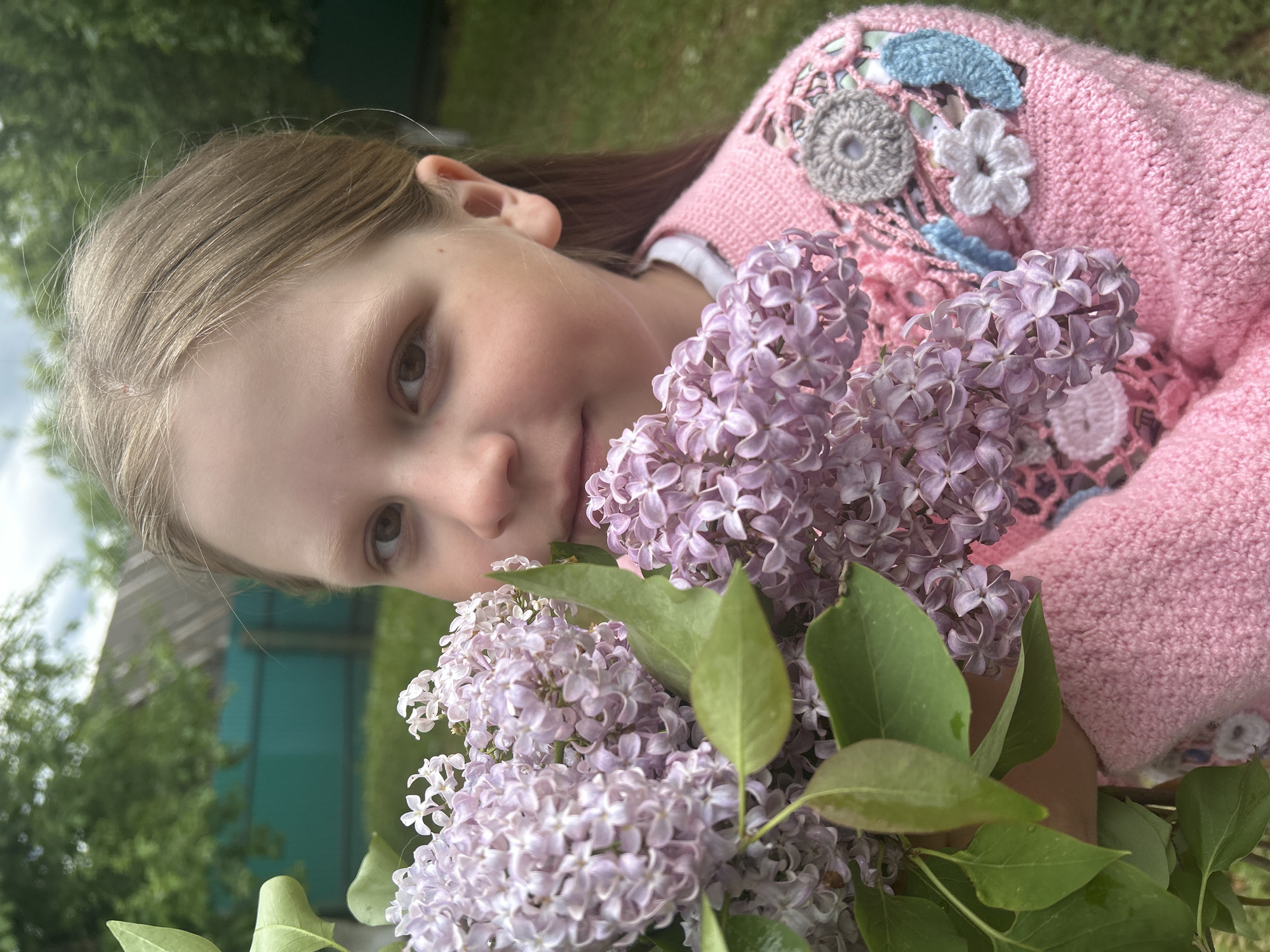 A young girl is featured in a close-up, partially obscured by a bouquet of purple lilacs with green leaves. She is wearing a pink knitted sweater decorated with crochet flowers and leaves. Her hair is blonde and parted to the side. The background shows green foliage and a turquoise structure. The location cannot be confirmed as Фоки, Russia from the visual information provided.