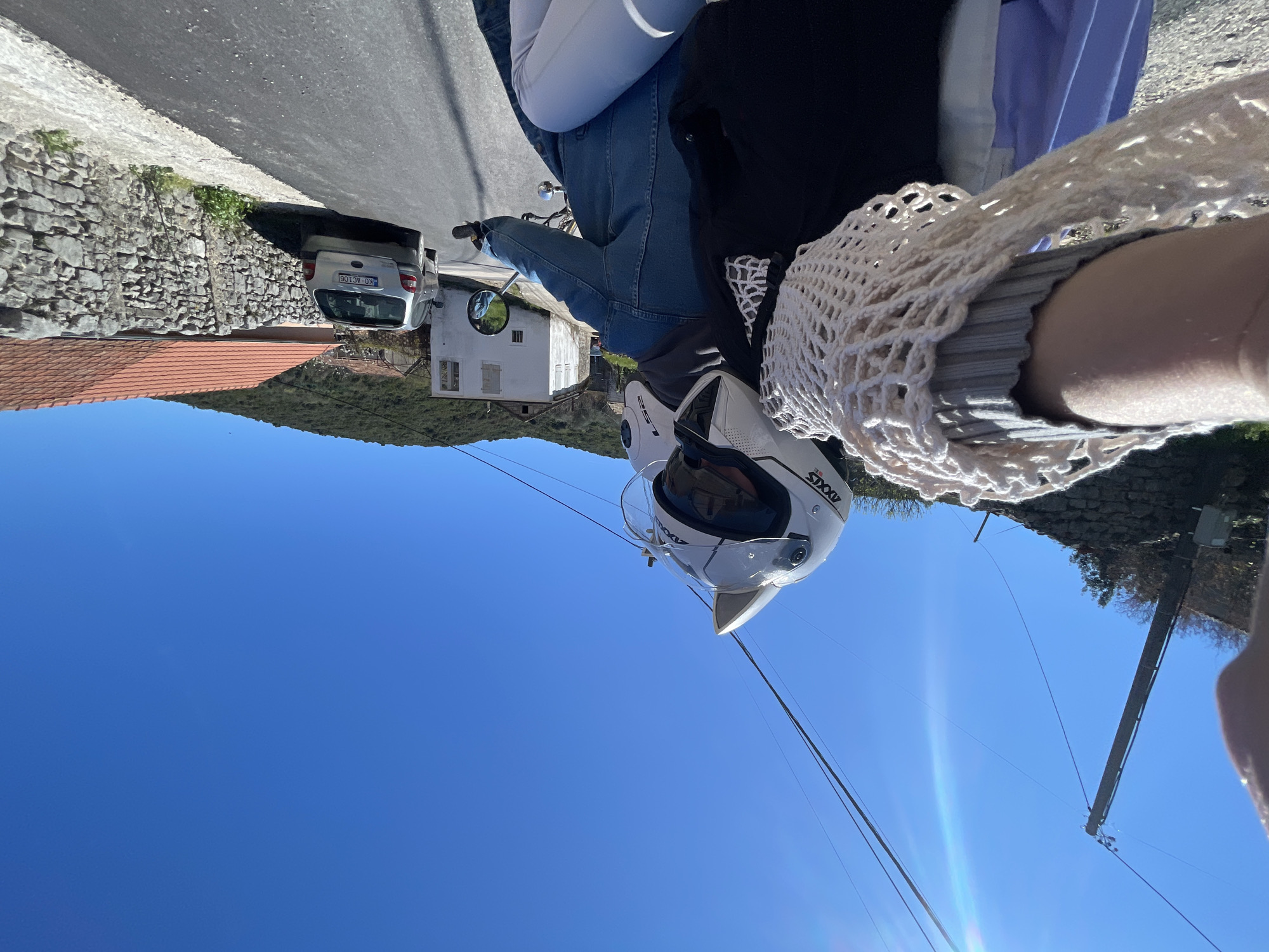 A rotated, selfie-style photo shows a person on a motorcycle, wearing a white Axxis helmet, riding on a road with a car parked nearby. The background includes a white house, trees, a brick wall, and a clear blue sky. Power lines stretch across the upper portion of the frame. The location appears to be Radanovići, Montenegro, based on the architecture and landscape.