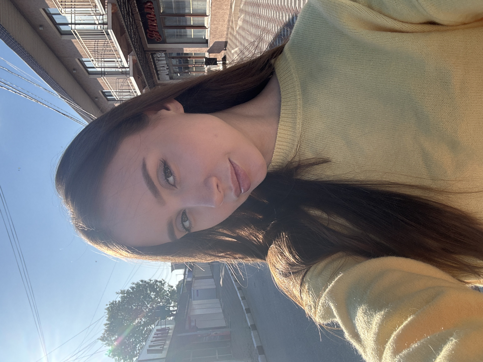 A woman is standing outside in a location near Береговой сельский совет, Ukraine. She is wearing a light yellow sweater. The sky is blue and sunny. In the background is a street with buildings and trees. There are power lines visible. The woman is holding the camera at an angle, taking a selfie.