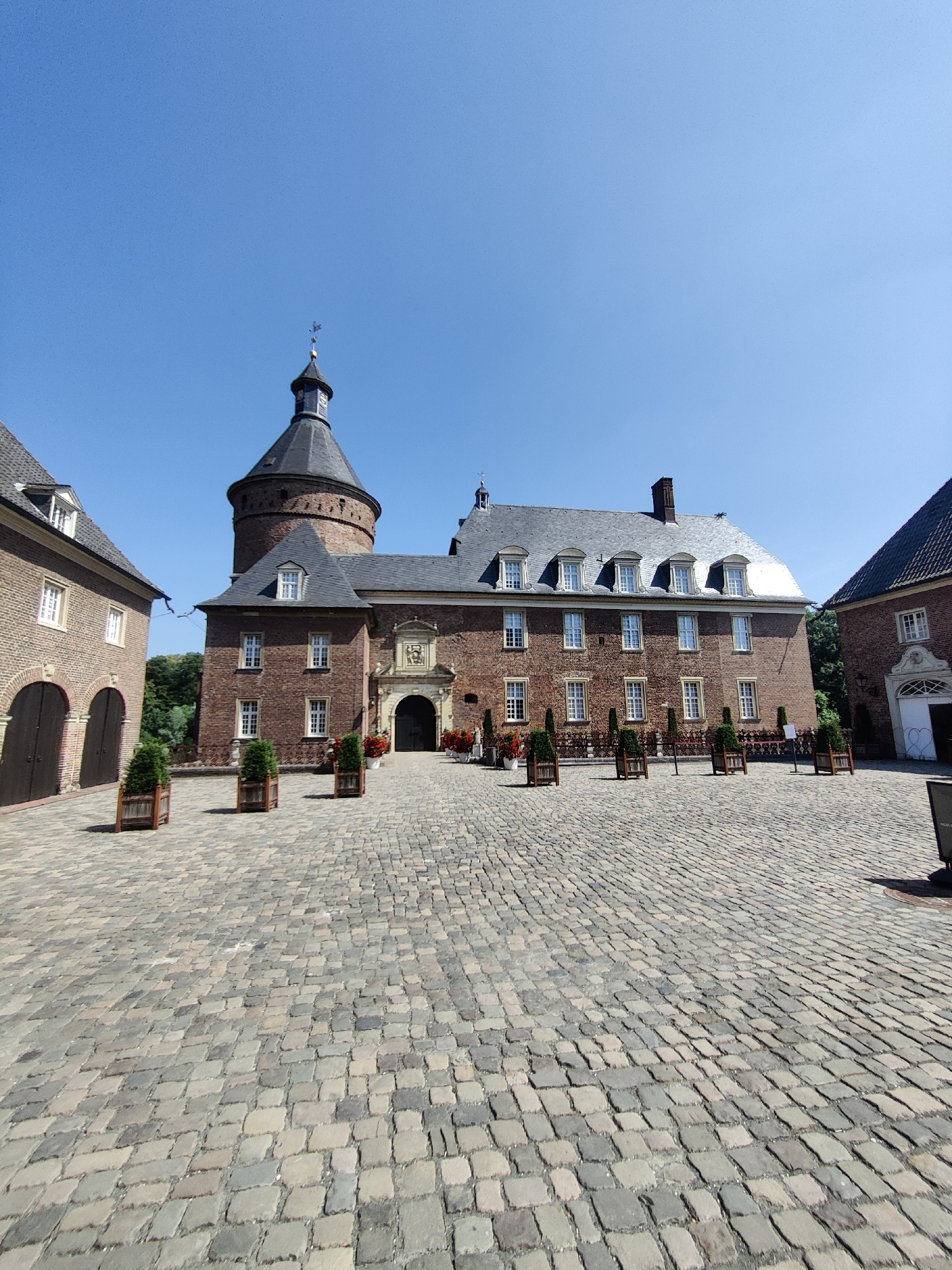 The image depicts the Anholt Water Castle (Wasserschloss Anholt) located in Isselburg, Germany, under a clear blue sky. The castle is a large, historic brick building with a prominent round tower topped with a dark spire and a weather vane. The main facade features multiple windows and a central arched entranceway with decorative elements.
In front of the castle is a wide, cobblestone courtyard. Several planters with small evergreen trees are strategically placed throughout the courtyard. To the left of the castle, a brick annex with arched doorways is visible. To the right, another part of the castle complex features a white facade with an arched entrance. The ground is paved with irregular-shaped cobblestones of varying shades of gray and beige, indicating a historic setting. The environment appears calm and sunny, with no people or animals visible in the frame. The overall impression is one of historical grandeur and architectural preservation.