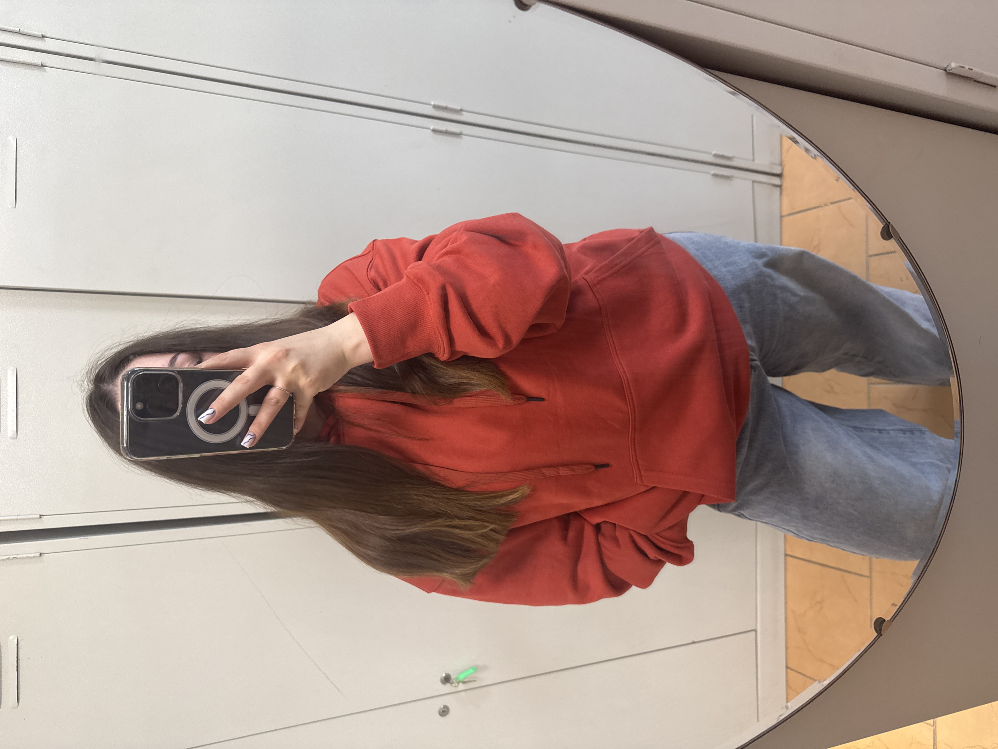 A person is taking a mirror selfie in what appears to be a locker room. The person is wearing a red hooded sweatshirt and light blue jeans. They are holding a smartphone with a magnetic ring on the back. In the background, there are white lockers and a tiled floor. The environment suggests a public facility like a gym, school, or community center.