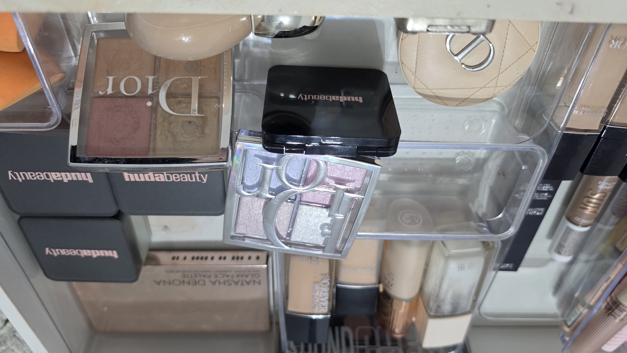 The image shows a drawer filled with makeup products. There are several items from Dior, including a blush palette, a face powder compact, and a few tubes of concealer or foundation. Some Huda Beauty products are also visible, including a blush compact and a few palettes. Additionally, there is a Natasha Denona Glam Face Palette. The makeup items are organized in clear plastic containers.