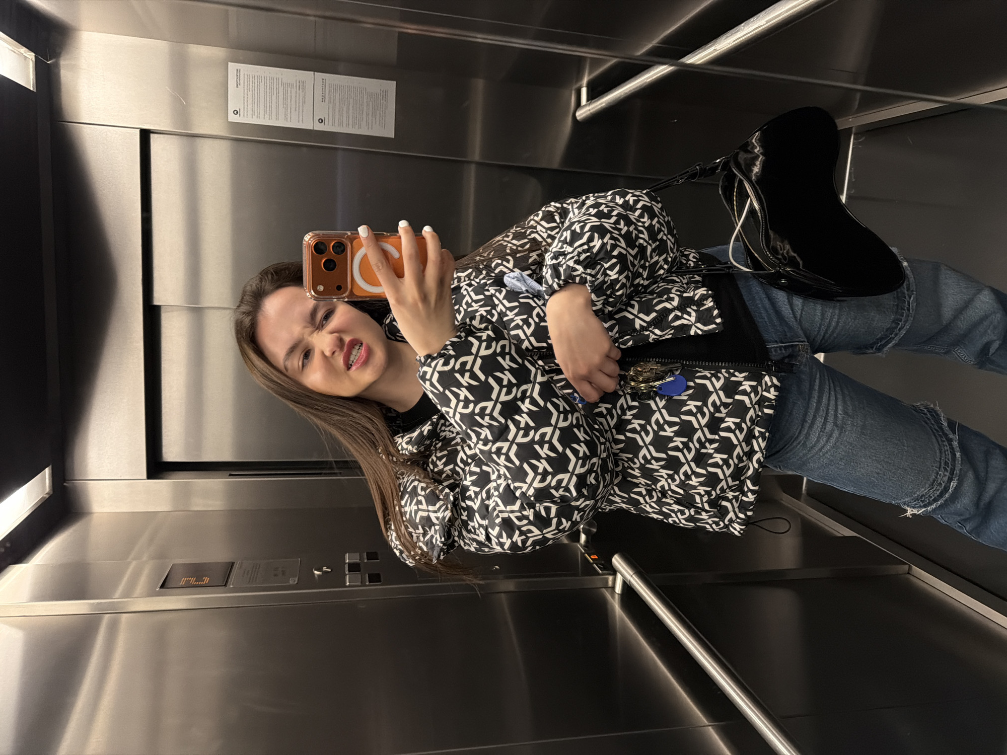 A young woman takes a selfie in an elevator. The elevator's interior is stainless steel. The woman wears a black and white patterned jacket, blue jeans, and black boots. She holds an orange smartphone with multiple lenses. The background includes elevator buttons and signage. This image was taken in Sofia, Bulgaria.