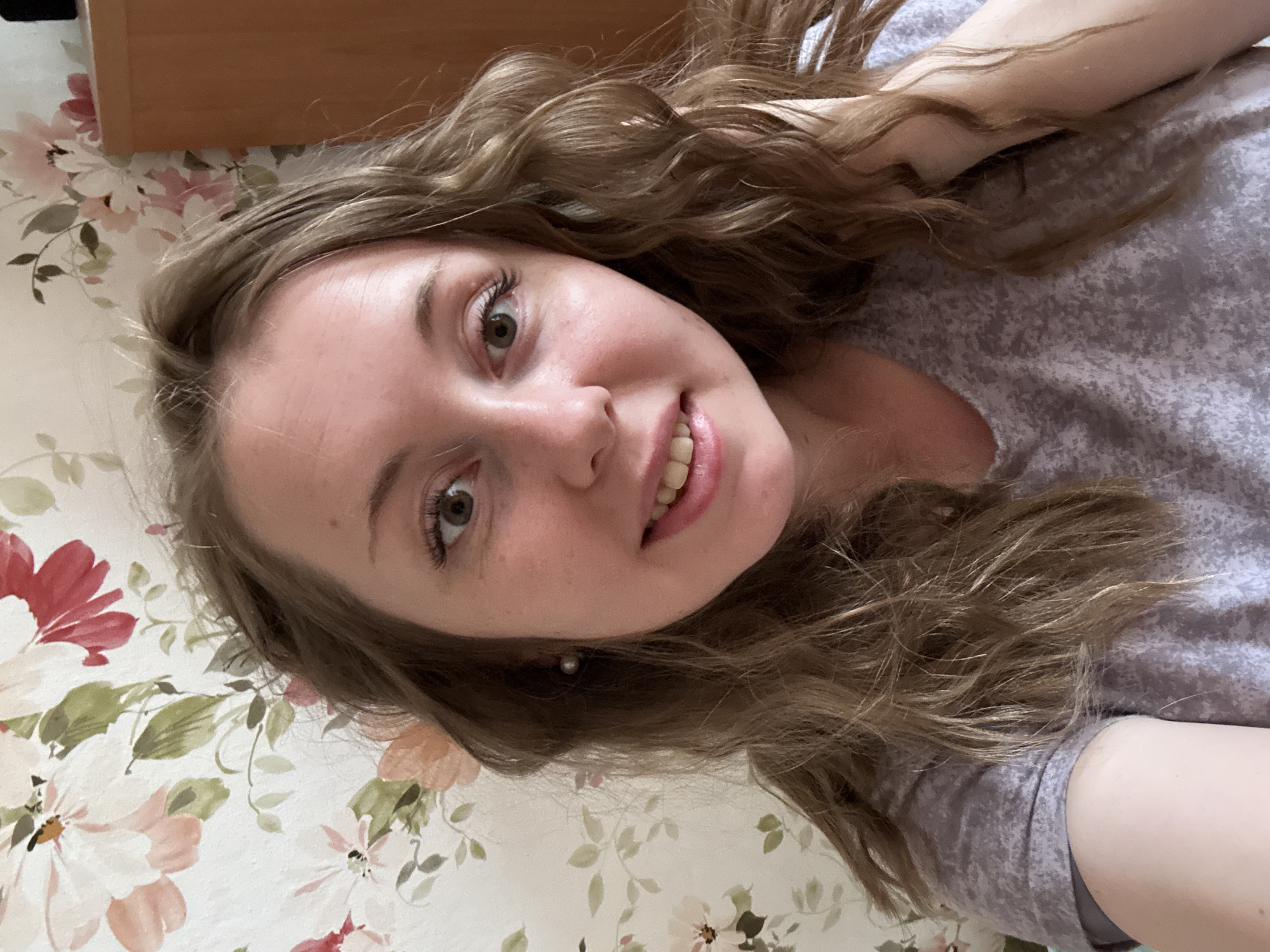 A young woman with wavy, light brown hair and fair skin is pictured in a close-up selfie. She is wearing a light gray shirt. Her eyes are light brown, and she is smiling slightly, revealing her teeth. She is wearing a small pearl earring.
The background behind the woman features a floral pattern with a white base and light red and pink flowers with green leaves. In the upper left corner, there is a section of a wooden surface.
The location of this scene is Unknown, Unknown.