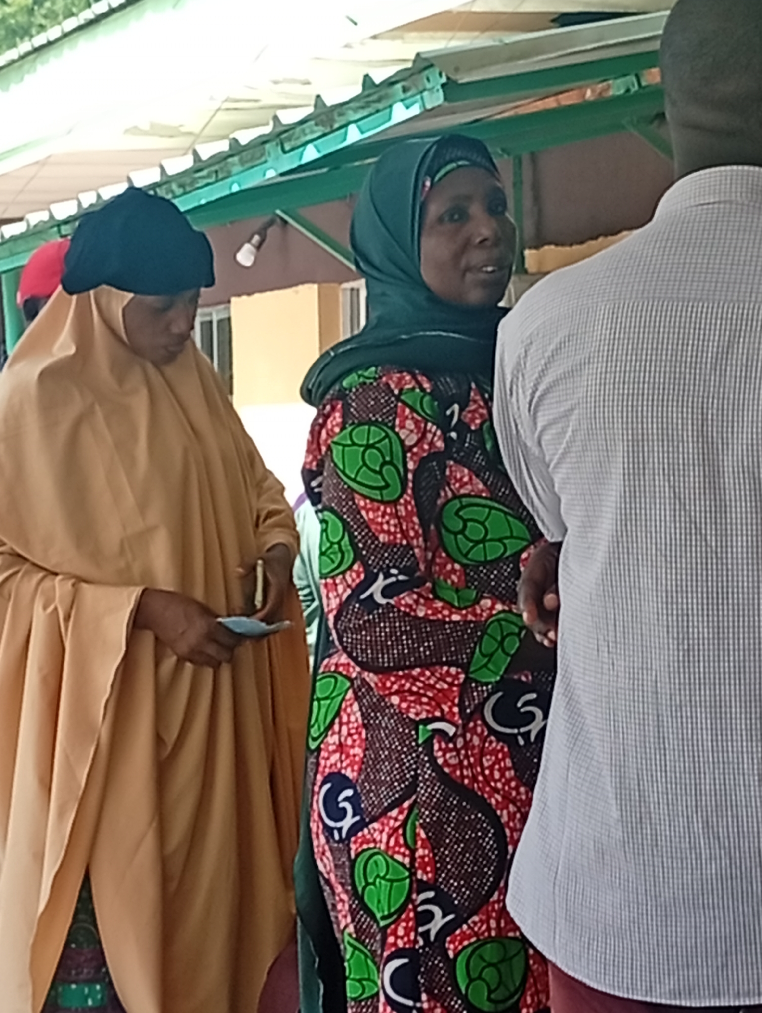 The image shows a group of people standing under a green-painted awning in what appears to be an outdoor market or gathering space in Gombe, Nigeria. The awning's metal frame is visible, as are some light fixtures attached to it. In the foreground, a woman wearing a tan hijab and a black head covering looks down as she holds what appears to be a small wad of cash or a ticket. To her right, another woman in a green hijab and a colorful patterned dress, featuring green circular motifs and red and white elements, is speaking. Her left hand is raised as if in mid-sentence. To the far right, a man wearing a light blue and white checkered shirt is seen from the back, facing away from the camera. The background consists of a building with yellow walls and some windows. The overall scene suggests a mundane daily activity in a Nigerian city.