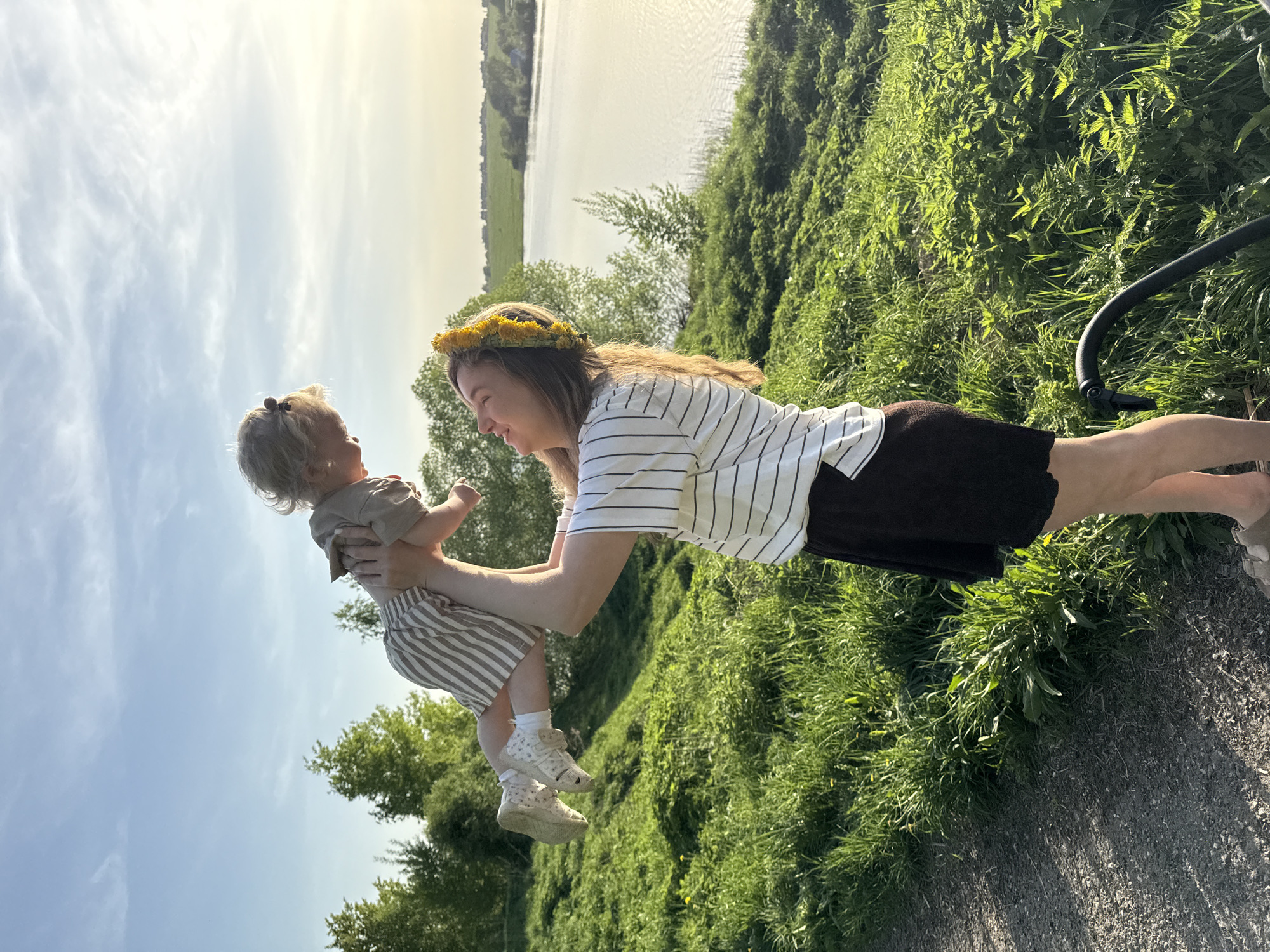 A woman wearing a sunflower crown and a striped shirt is holding a toddler in the air. The toddler is wearing a brown shirt and striped shorts. They are standing in a grassy area next to a path. In the background is a body of water with trees on the opposite bank. The scene is likely in Vladimir, Russia.