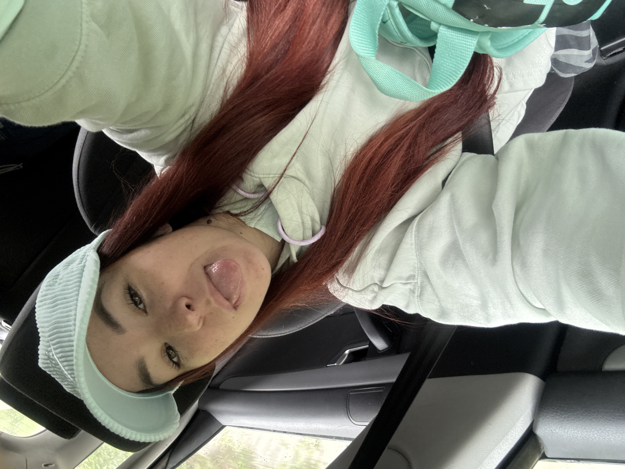 A young woman with red hair and freckles is taking a selfie inside a car. She is wearing a light blue hooded sweatshirt and a light blue visor. She is sticking her tongue out. A black seat and seatbelt are visible, as is a light blue backpack. The location in the city of Novosellë, Kosovo is not discernible.