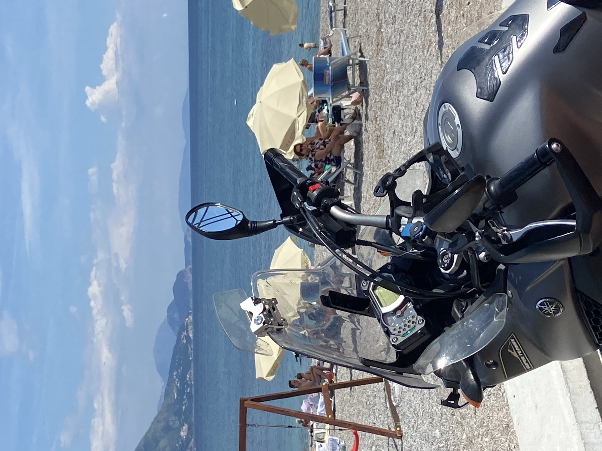Here's a description of the image:
The image is taken from the perspective of a motorcycle parked on what appears to be a concrete surface. The foreground is dominated by the motorcycle's handlebars, instrument panel, and part of the fuel tank.
In the midground, a beach scene unfolds. There are several beach umbrellas and people lounging in chairs or on towels. Some appear to be sunbathing.
The background reveals a body of water, likely a sea or lake, with landmasses visible in the distance. The sky is blue with scattered clouds.
Based on the prompt's provided information, the location is likely the city of Prijevor in Montenegro, specifically a beach area.