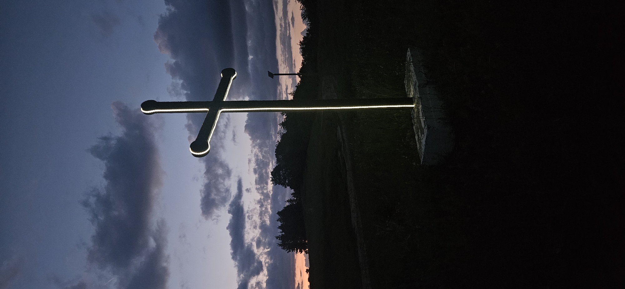 An outdoor long-shot photograph taken at dusk or dawn in Gutsal, Bulgaria, centers on a large, dark, illuminated cross in the mid-foreground, slightly right of the vertical axis. The cross features thick, tubular construction with rounded ends and is outlined by a bright, continuous white light source, likely an LED strip. It stands on a rectangular concrete base. To the right and further back, a slender dark pole with a small solar panel mounted atop is visible. The foreground consists of dark, uncultivated ground covered in low vegetation. The midground features a dark silhouette of a tree line extending across the horizon, with what appears to be a dirt path or track curving through the grassy field to the left. The sky exhibits a gradient of colors, transitioning from a deep purplish-blue at the zenith to a muted reddish-orange glow along the left horizon. Scattered, dark clouds are present throughout the sky. No individuals, animals, or specific actions are depicted.
