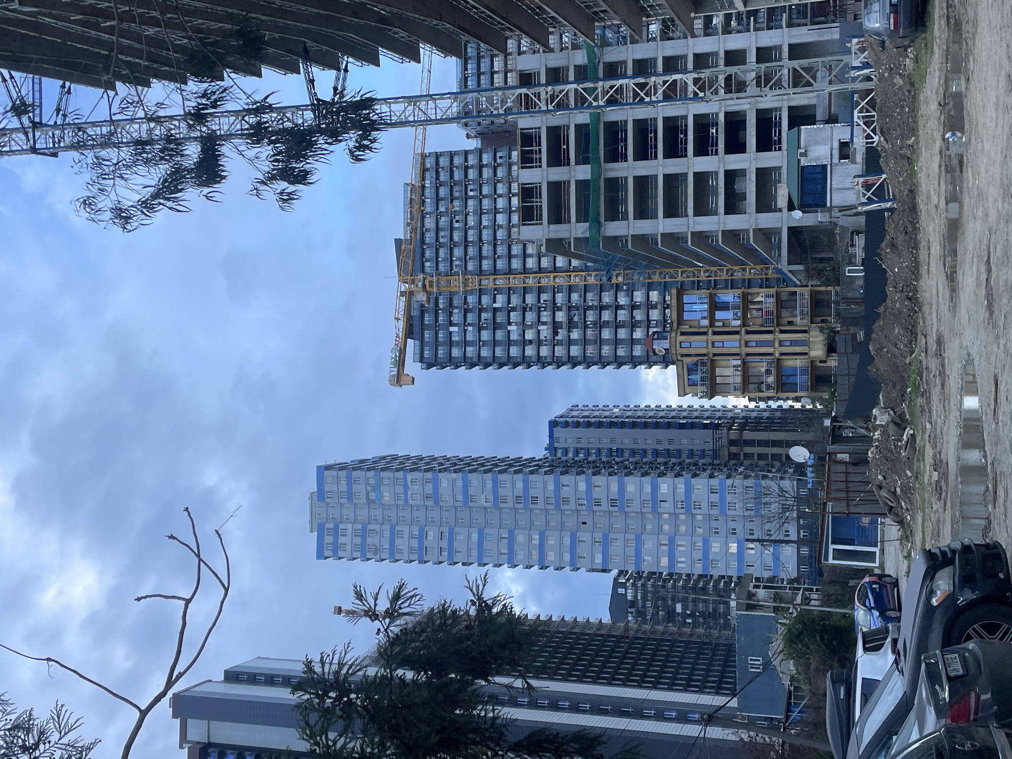 The image depicts a construction site in Batumi, Georgia. Multiple high-rise buildings are visible, some under construction with cranes present. Buildings show exposed concrete structures and partially completed facades. There is a collection of parked cars in the foreground. The ground appears to be unpaved and muddy. The sky is overcast.