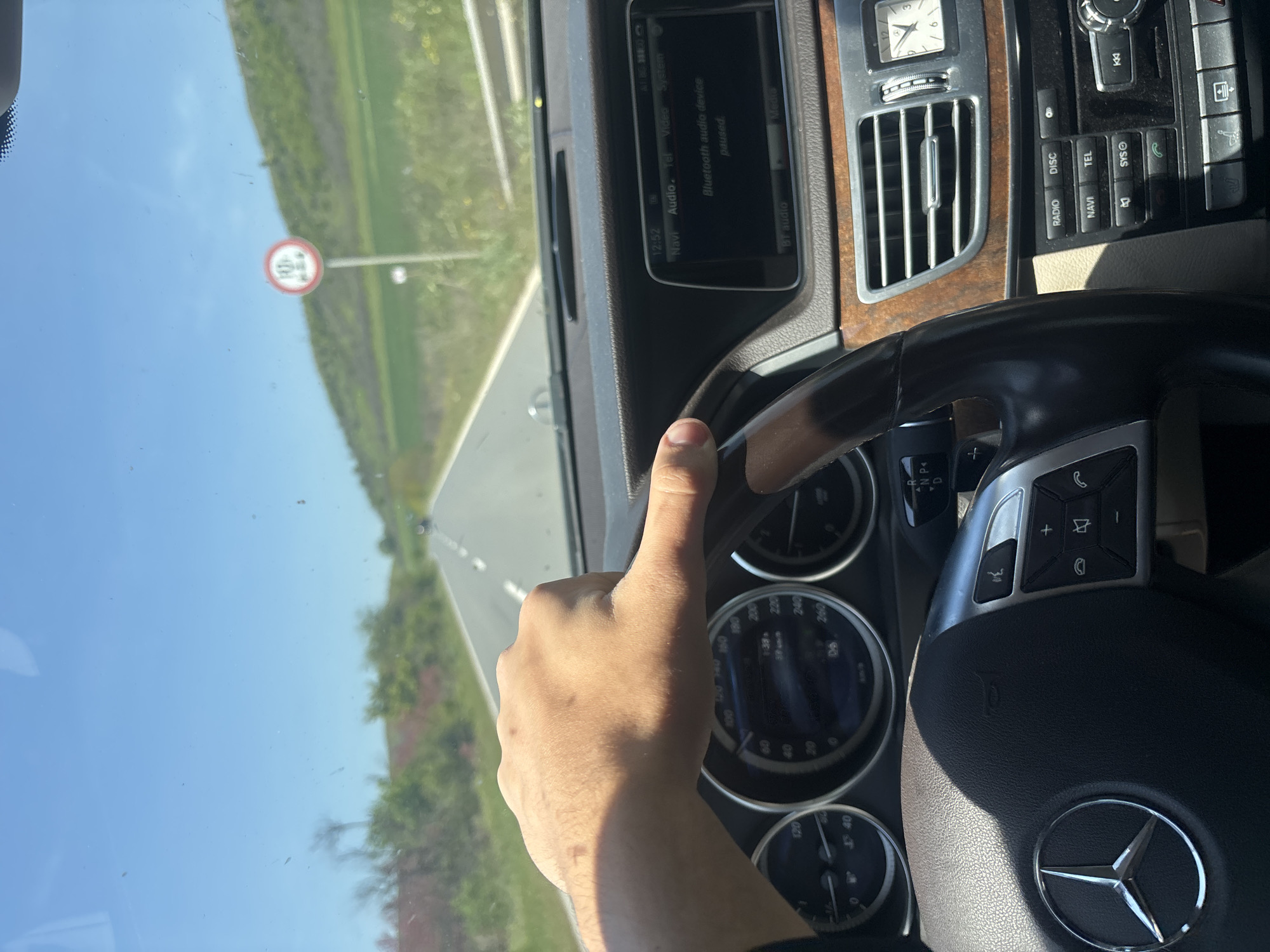 The image shows a person driving a Mercedes-Benz sedan on a highway with a speed limit sign of 40 km/h visible in the distance. The interior of the car is visible, including the steering wheel, dashboard, infotainment system displaying "Bluetooth audio device paused", and instrument cluster. The driver's hand is visible, holding the steering wheel. The location is likely in Bulgaria due to the Cyrillic text on the speed limit sign. The specific city of Bata cannot be confirmed from the image.