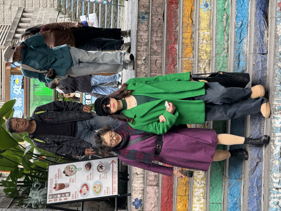 The image depicts an outdoor, multi-level pedestrian staircase in Yuzhong District, China. The stone stairs are painted with numerous horizontal stripes in vibrant colors, including red, orange, yellow, green, and blue, with some steps featuring small floral patterns.
In the foreground, two women are descending the colorful steps, arm-in-arm. The woman on the left, appearing middle-aged, wears glasses, a purple knee-length coat cinched with a black belt, a red and black plaid scarf, beige tights, and black lace-up boots. She holds a dark smartphone in her right hand. The woman on the right, also appearing middle-aged, wears a bright green overcoat, a black baseball cap, large black square sunglasses, dark trousers, and light brown slipper-style shoes. She carries a black quilted handbag on her left arm and holds a small white rectangular object in her right hand.
Behind them, further up the stairs, several other individuals are present. An older man stands centrally, wearing a grey and black patterned hooded jacket with bright yellow accents, a black t-shirt, and blue jeans. To his right, a woman in a light teal belted jacket, light grey pants, and a light-colored cap is visible, seemingly holding a phone. Another woman in a brown top and dark pants stands to her right. Further back, additional indistinct figures are visible on the ascending steps.
To the immediate left of the stairs, a white A-frame menu board stands, titled "DELICIOUS" and displaying images and descriptions of various food and drink items, labeled "TOP1," "TOP2," etc. Green foliage is visible in the upper left portion of the image, and building facades with a blue sign featuring Chinese characters are present in the background.