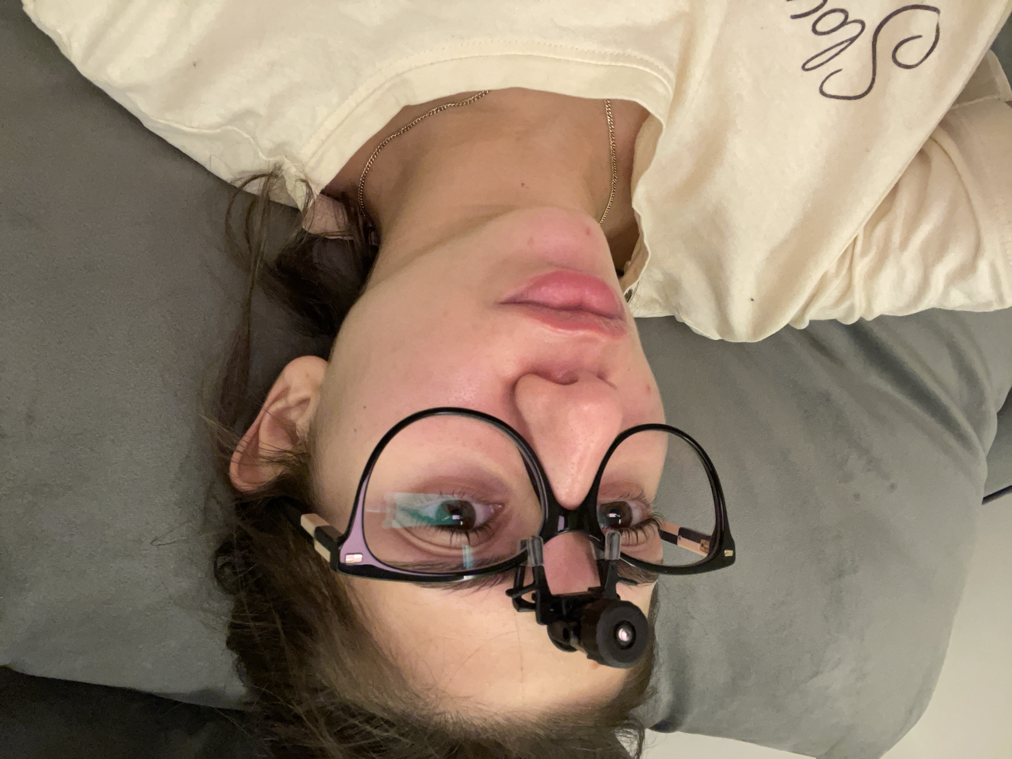 A woman is lying on a gray surface, possibly a bed or couch, wearing glasses with a head-mounted magnifier attached. She is wearing a light-colored t-shirt with dark lettering. A gold chain is visible around her neck. Her face is facing upwards toward the camera. The image was not taken in Sofia, Bulgaria.