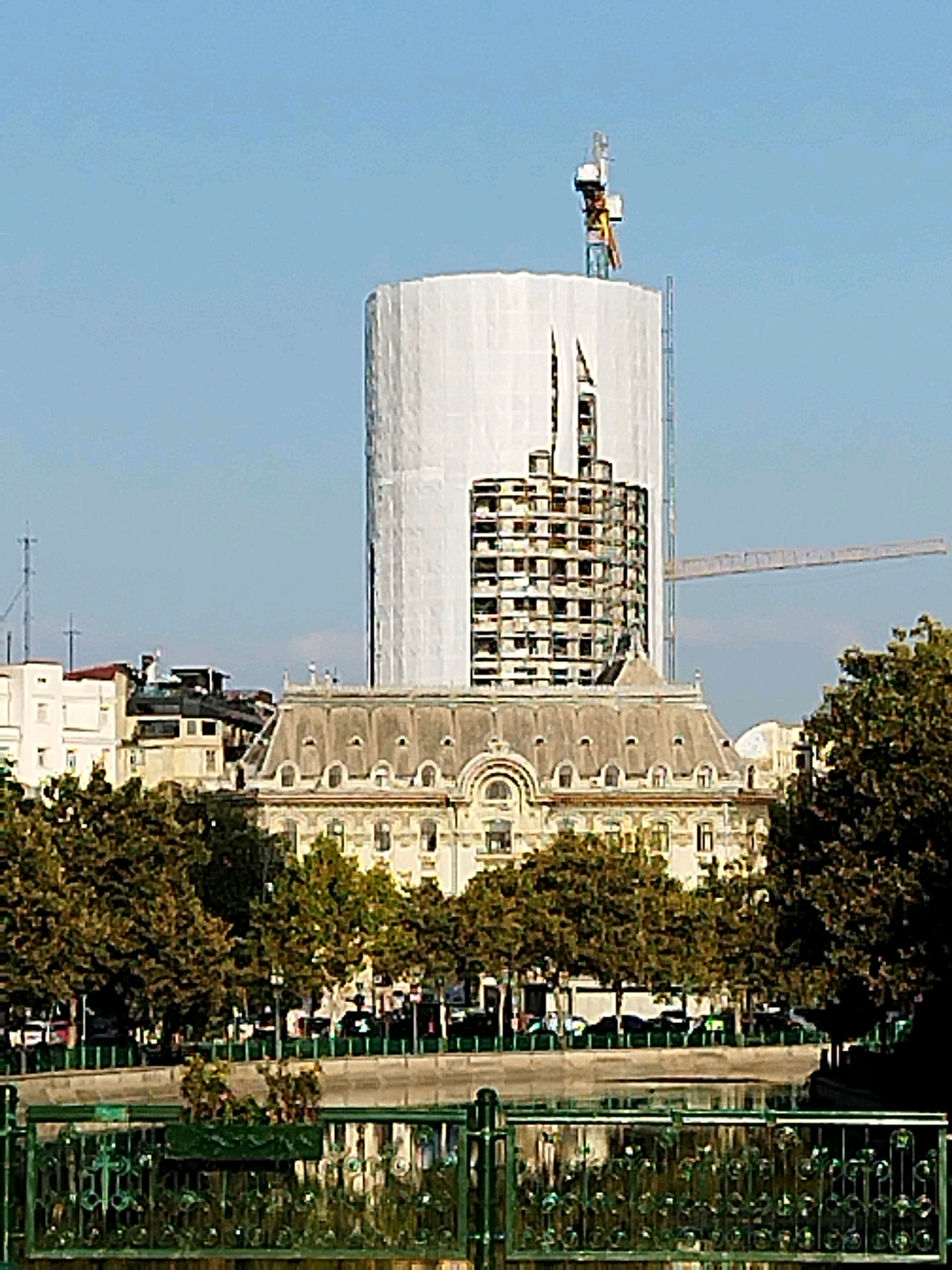In Bucharest, Romania, a cylindrical building under construction is partially covered in white sheeting, with visible structural elements and scaffolding. A construction crane is positioned at the top of the building. In front of the unfinished structure, an ornate, mansard-roofed building stands, partially obscured by lush green trees. The scene also includes a body of water with reflections and a decorative green fence in the foreground, suggesting a park or riverside setting within the city. The sky is clear and blue.