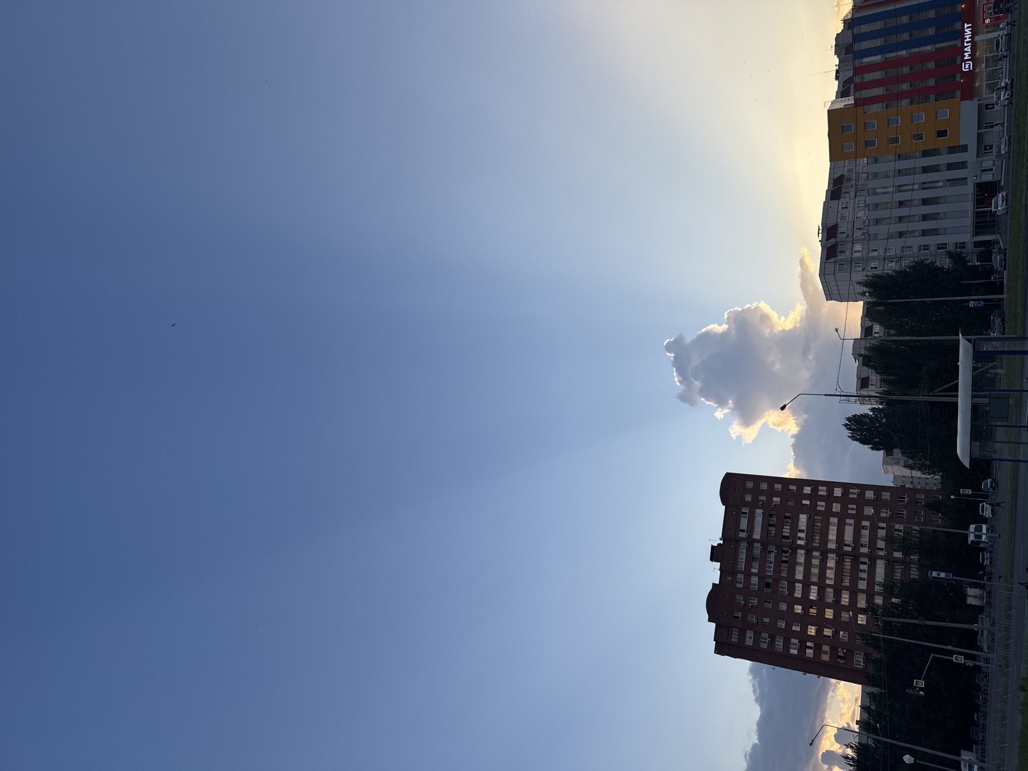 This is a photograph of a city skyline, likely Tolyatti, Russia, taken during sunset. The sky is a clear blue with rays of light emanating from behind a large, dramatic cloud formation. In the foreground, a tall, reddish-brown brick apartment building dominates the left side of the frame, its windows reflecting the setting sun. To the right, a modern building with a colorful facade, featuring sections of yellow, blue, and red, is visible. The ground level of this building displays the "Магнит" logo, indicating it is a supermarket. Trees line the street in front of the buildings, and streetlights and traffic lights are present. A bus stop shelter is visible in the lower right corner. Cars are seen on the street, and a traffic light is on the left side of the image. The overall impression is one of urban life transitioning into evening.
