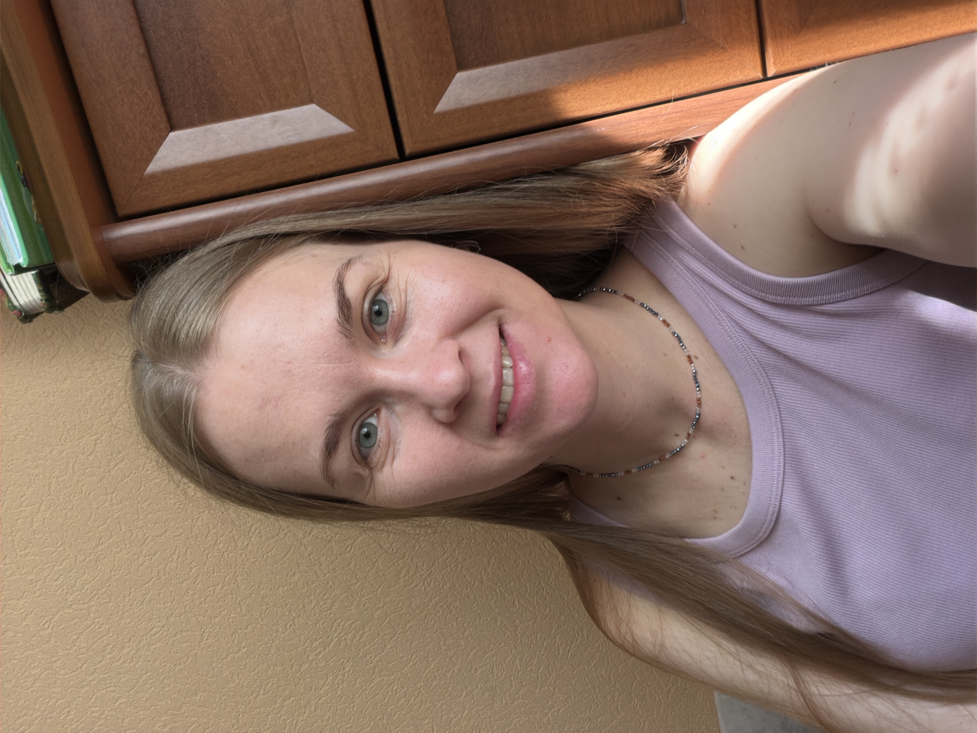 A woman with blonde hair and blue eyes smiles at the camera. She is wearing a lavender tank top and a beaded necklace. She is standing in front of a wall with a textured pattern. To her right is a wooden dresser with several drawers. Sunlight is casting a shadow on her arm and the side of the dresser. The image appears to be taken indoors, in what could be a bedroom. There is no information in the image to suggest it is located in Khabarovsk, Russia.